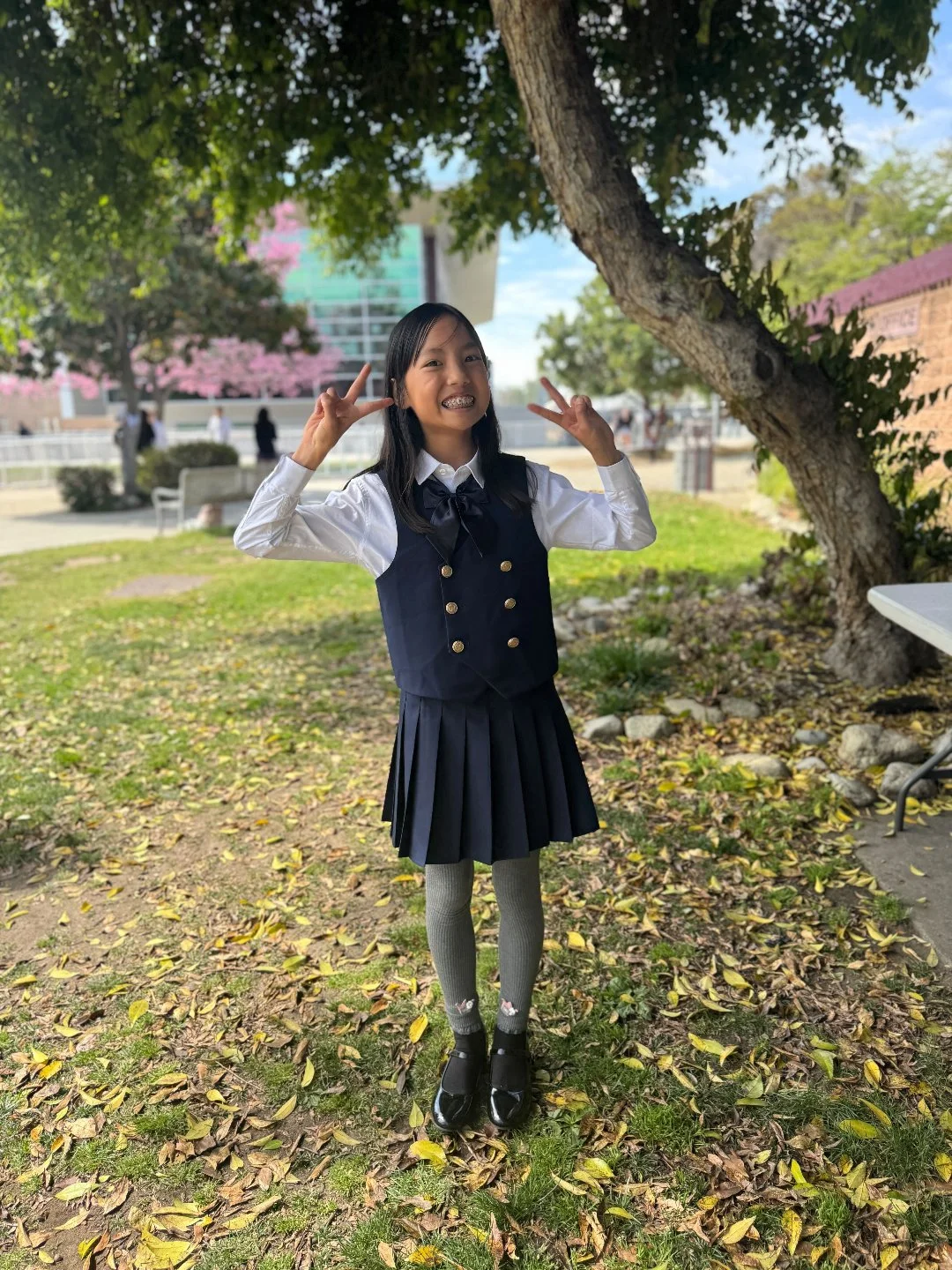 A young girl wearing a navy blue school uniform with a pleated skirt, gray knee-high socks, black shoes, and a white shirt with a navy blue tie, standing outdoors under a large tree, smiling and making peace signs with both hands on a sunny day.