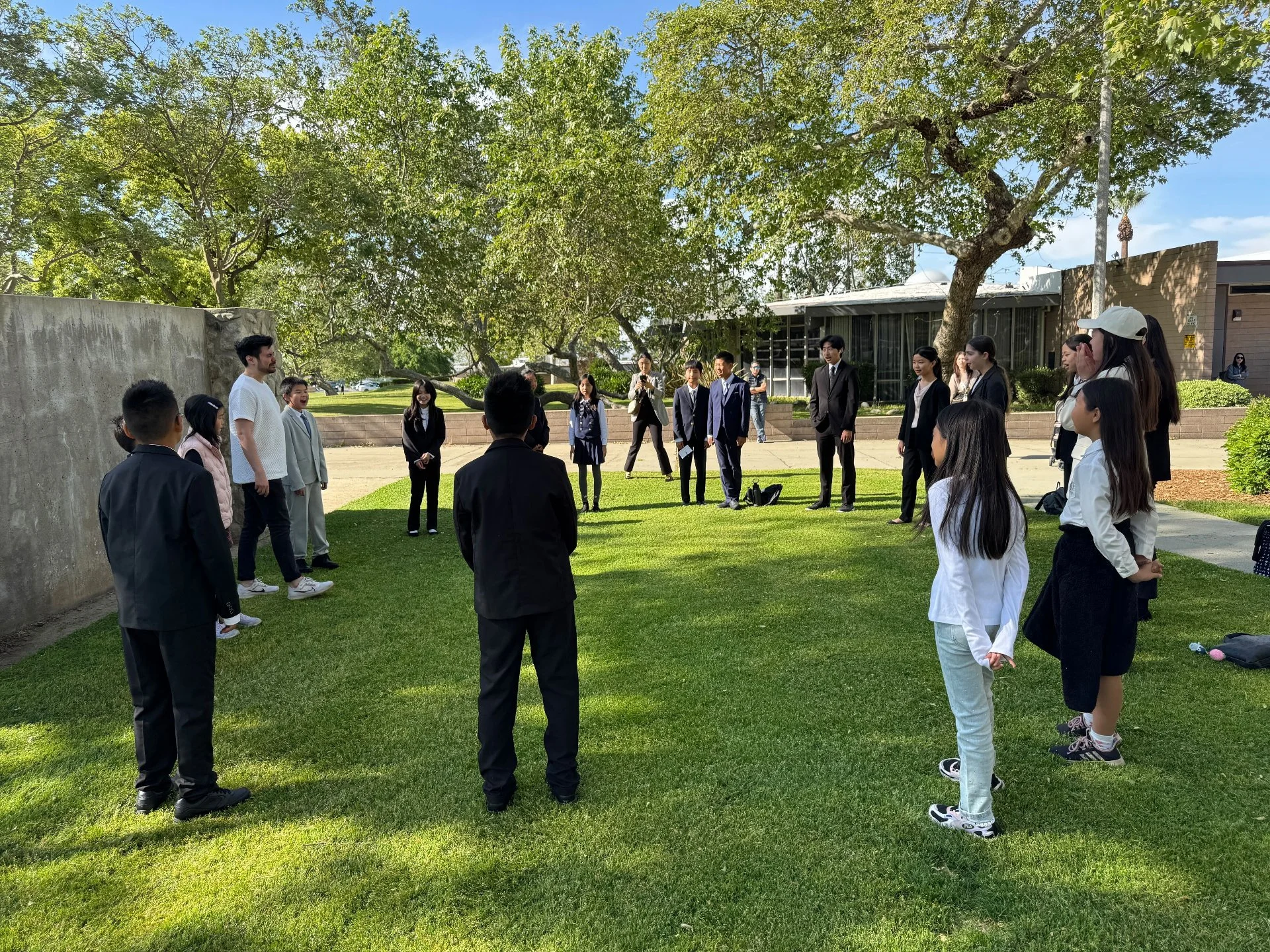 A group of people gathering outdoors in a circle on green grass, possibly participating in a team-building or outdoor activity, with trees and a building in the background.