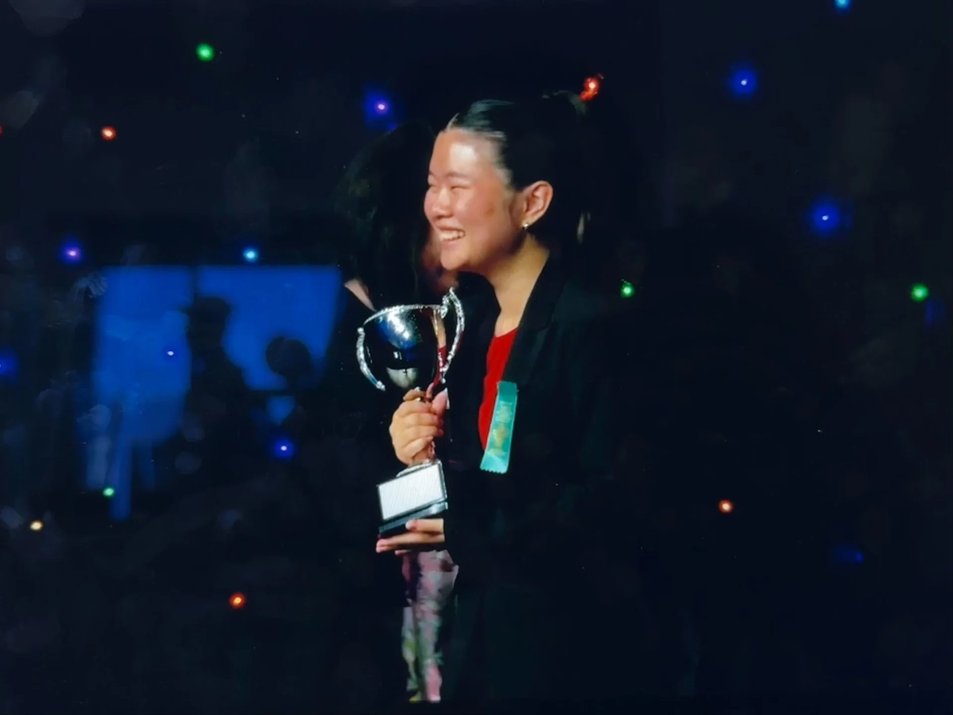 A woman smiling and holding a trophy, standing against a background with colorful lights and a blue screen.
