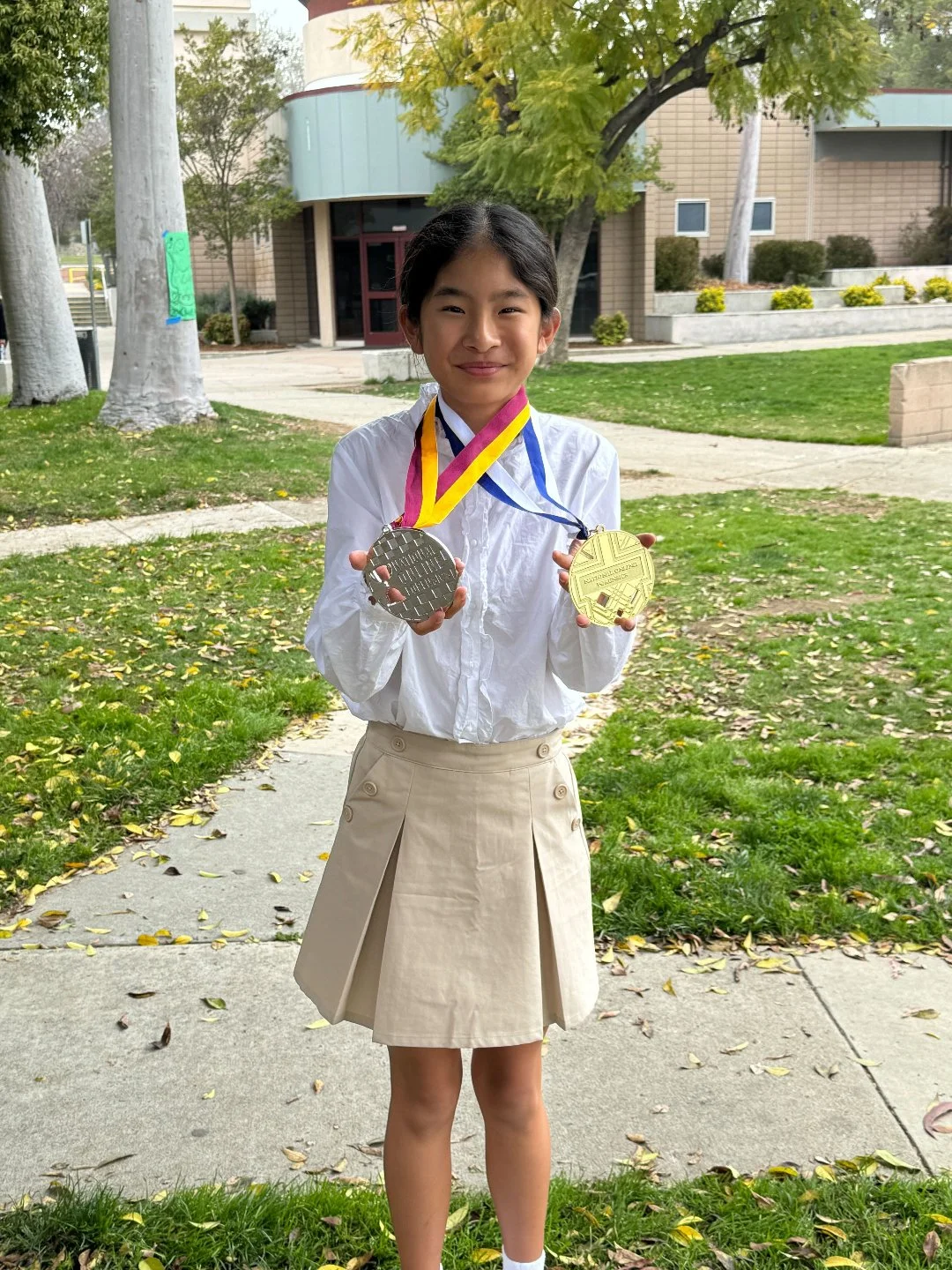 A young girl holding two medals, one silver and one gold, outdoors on a sidewalk with grass and trees in the background.