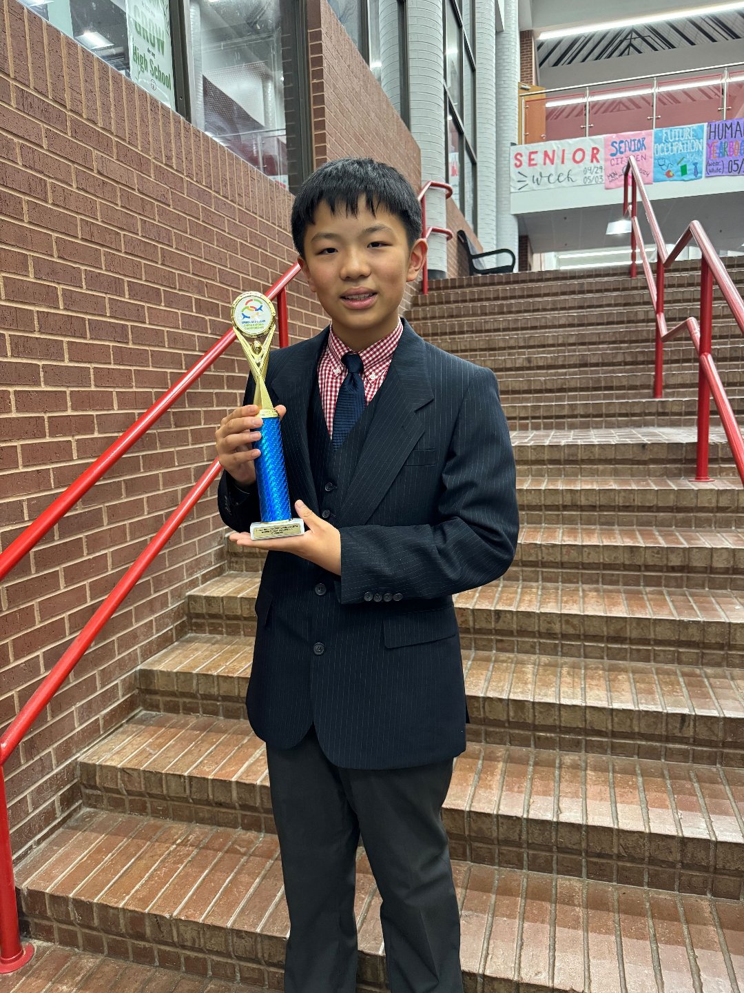 A young boy in a dark suit and tie, holding a trophy, standing on brick stairs with red railing, in an indoor school setting.