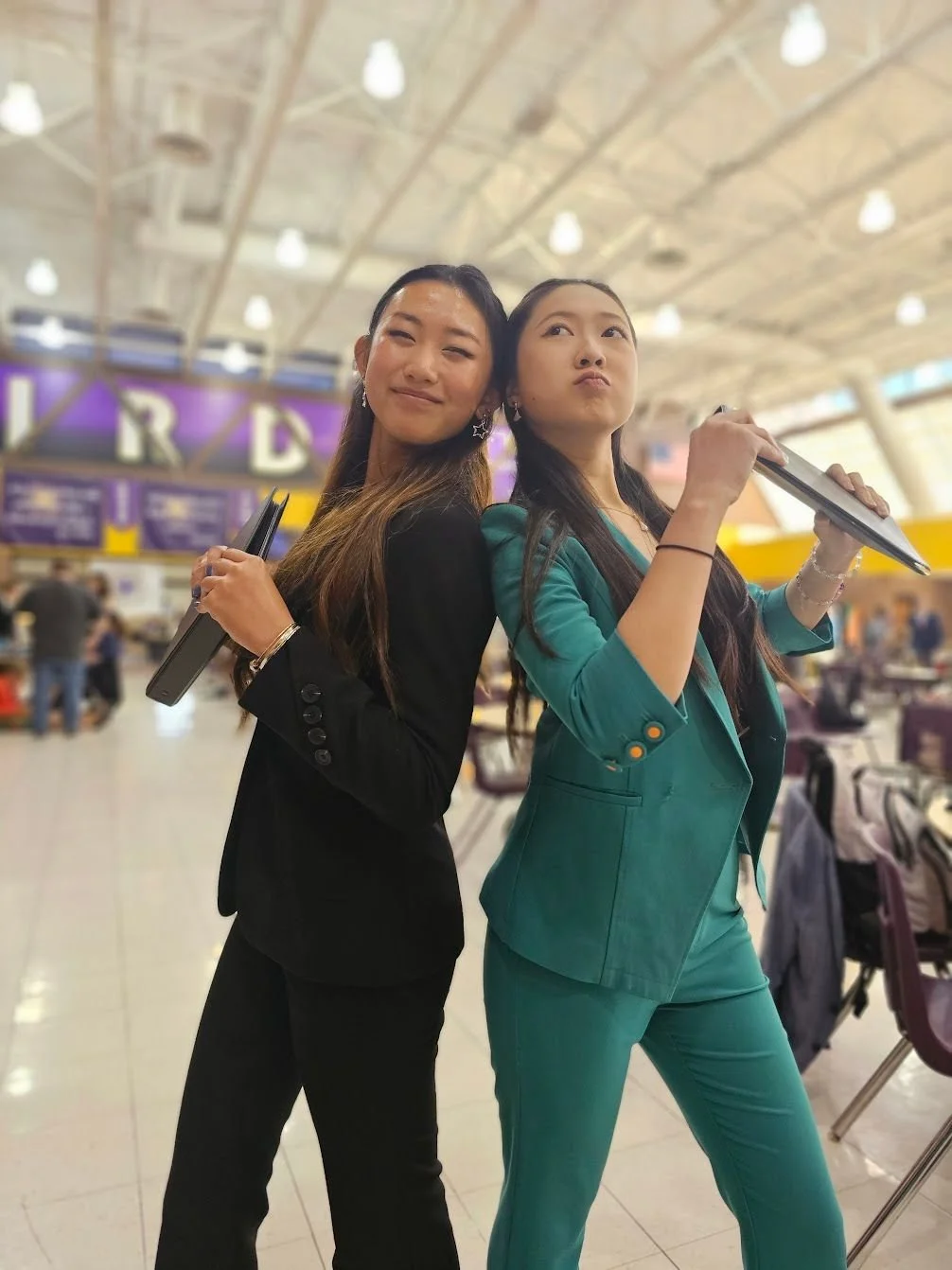Two women stand back to back in an airport terminal, holding boarding passes, with the letter 'R' visible on a signboard in the background.