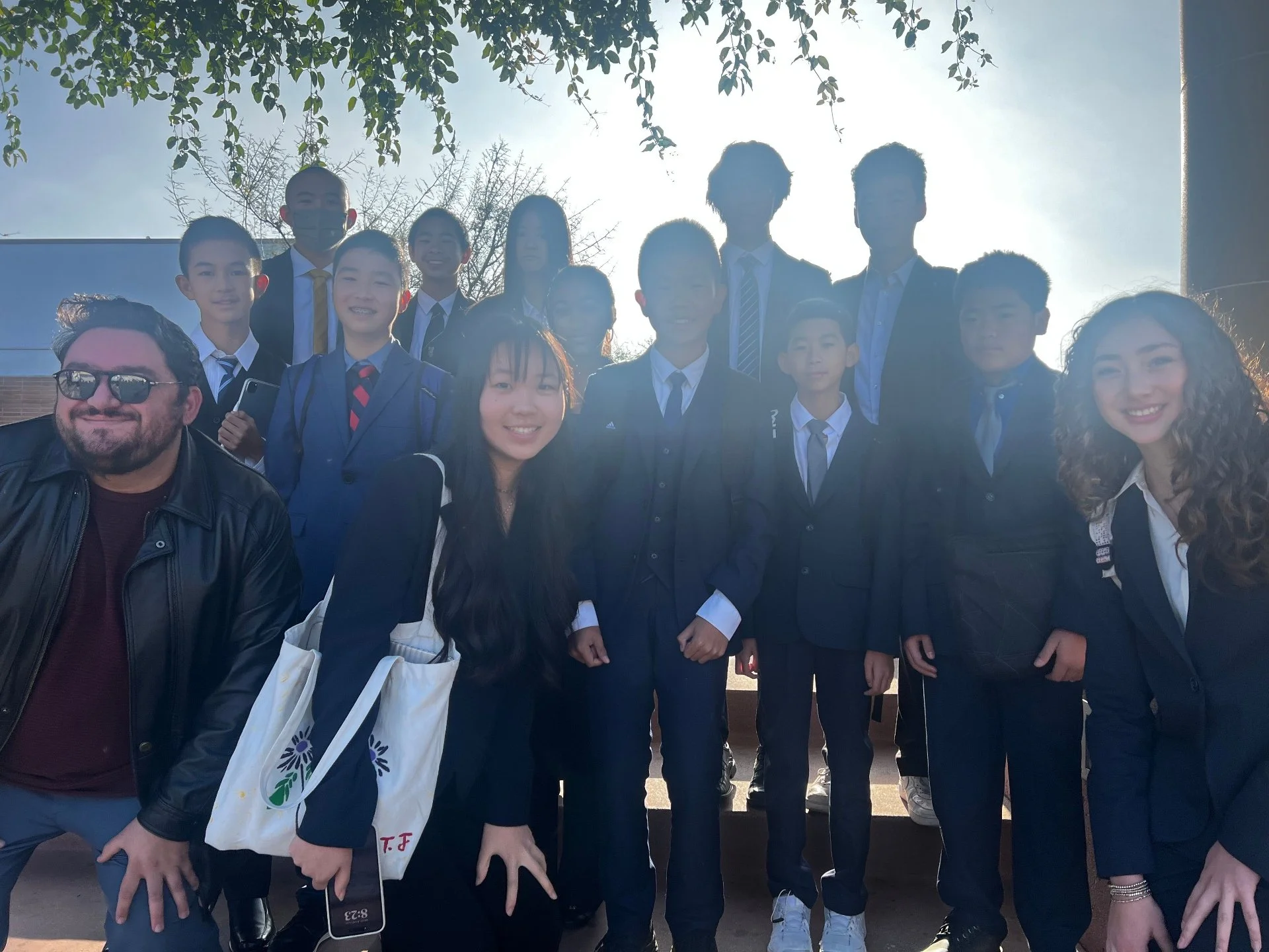 Group of students and adults posing outdoors, some in school uniforms, with a bright sky and trees in the background.