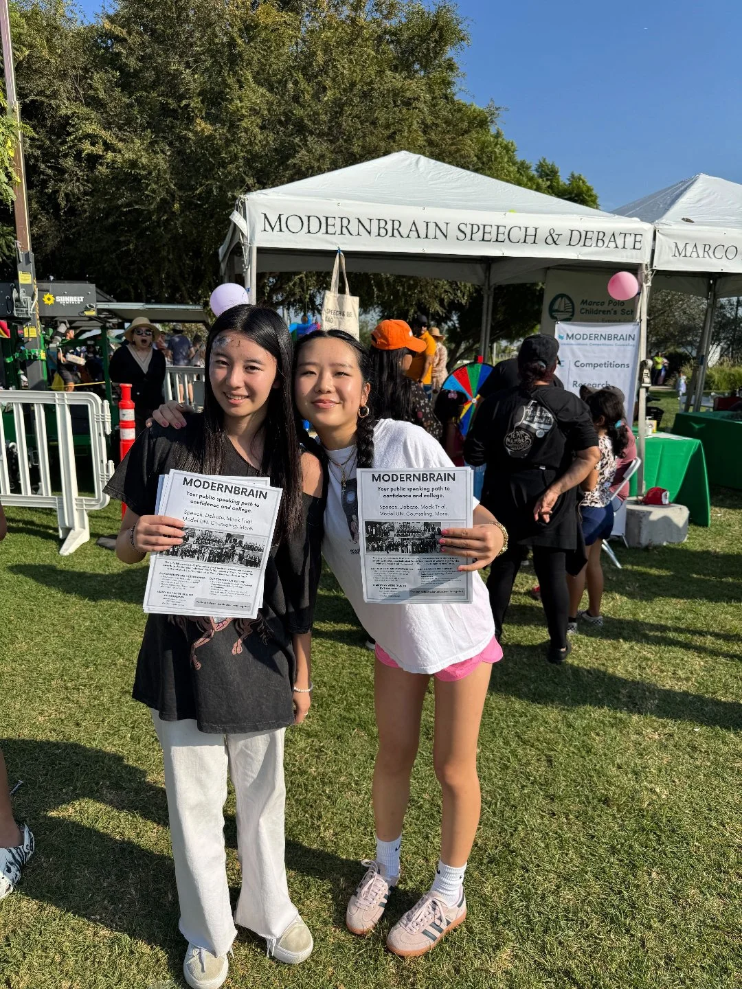 Two young women smiling and holding informational flyers at an outdoor event, standing in front of a tent labeled 'Modern Brain Speech & Debate'.