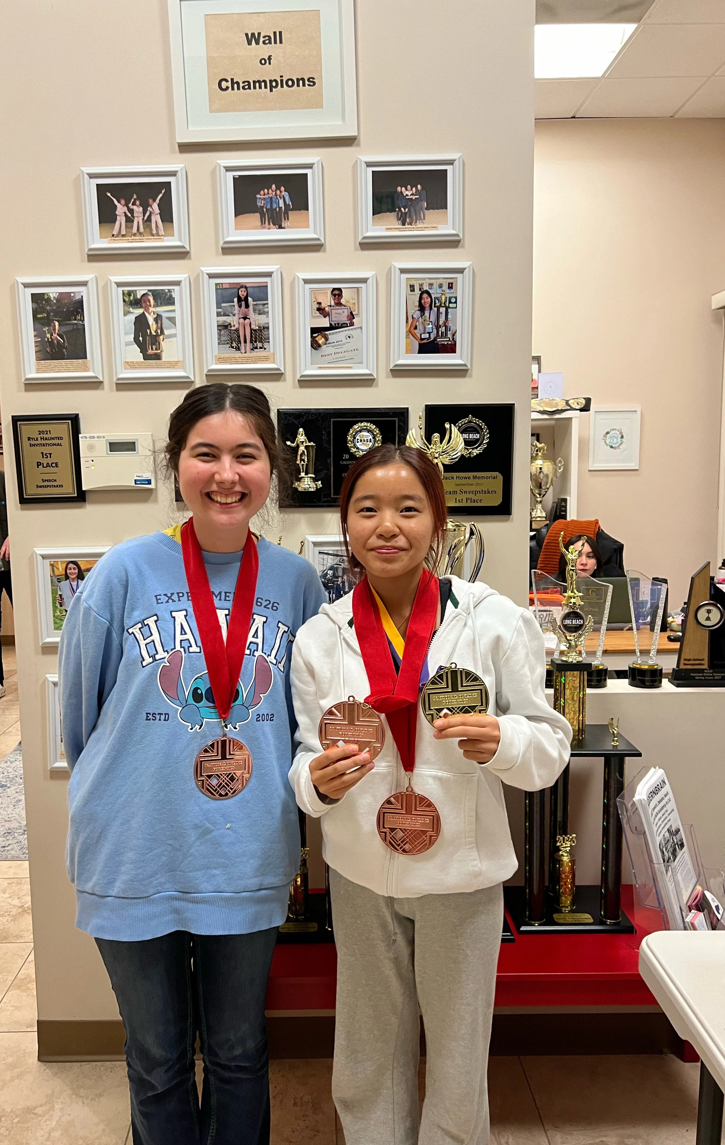 Two young women displaying medals, standing in front of a wall with trophies, photos, and plaques. One is smiling, wearing a blue hoodie, and the other has a neutral expression, wearing a white hoodie. The medals are bronze and gold. The photo appear