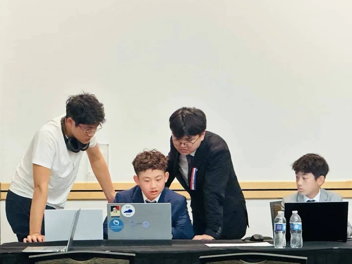 Four young men sitting at a table with laptops and water bottles, engaged in a discussion or working together, two standing and two sitting, in a professional setting.