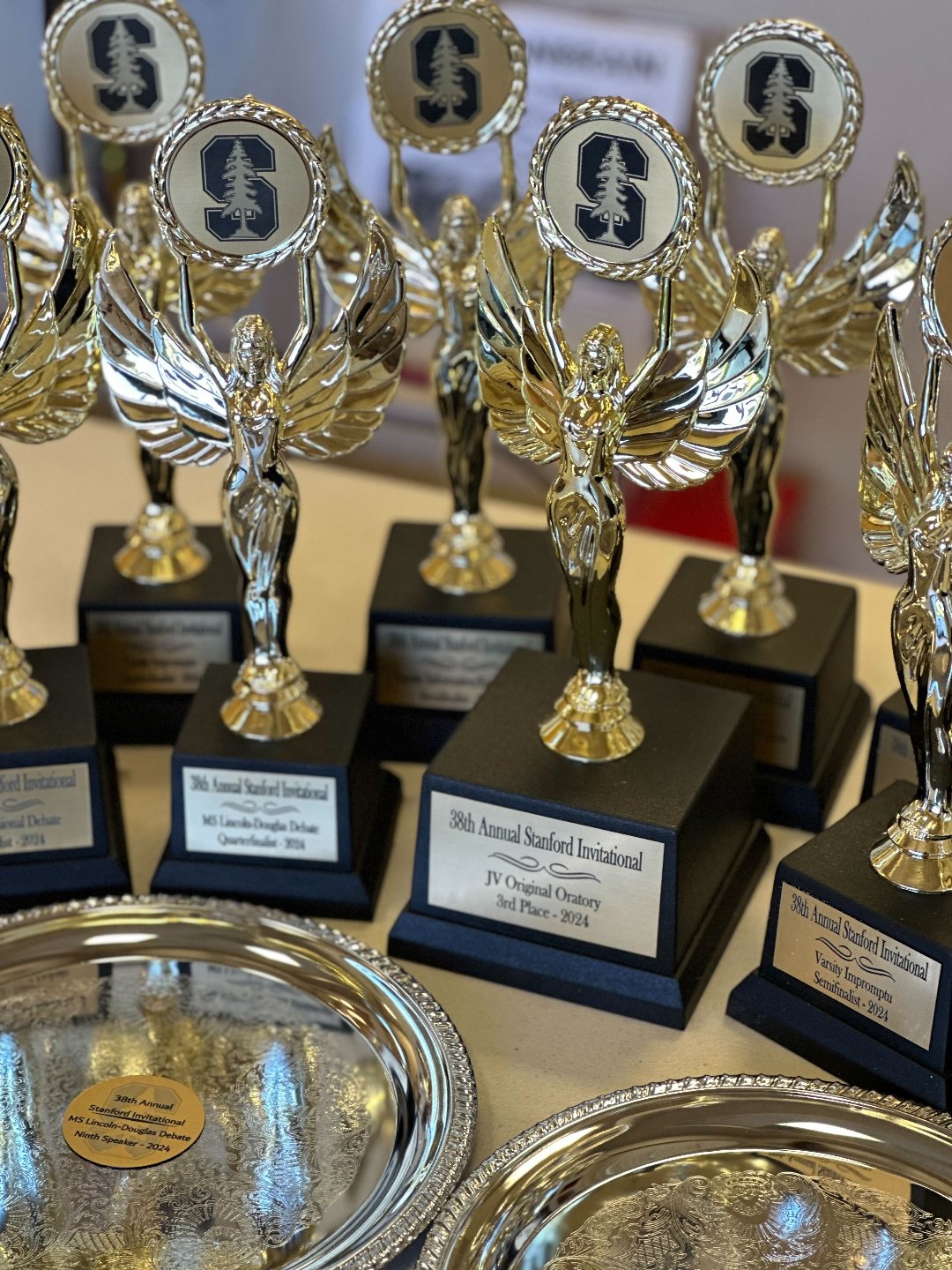 Several gold and silver trophies with winged female figures holding plaques, displayed on black bases, with some plaques reading Stanford Invitational awards from 2014 to 2024.