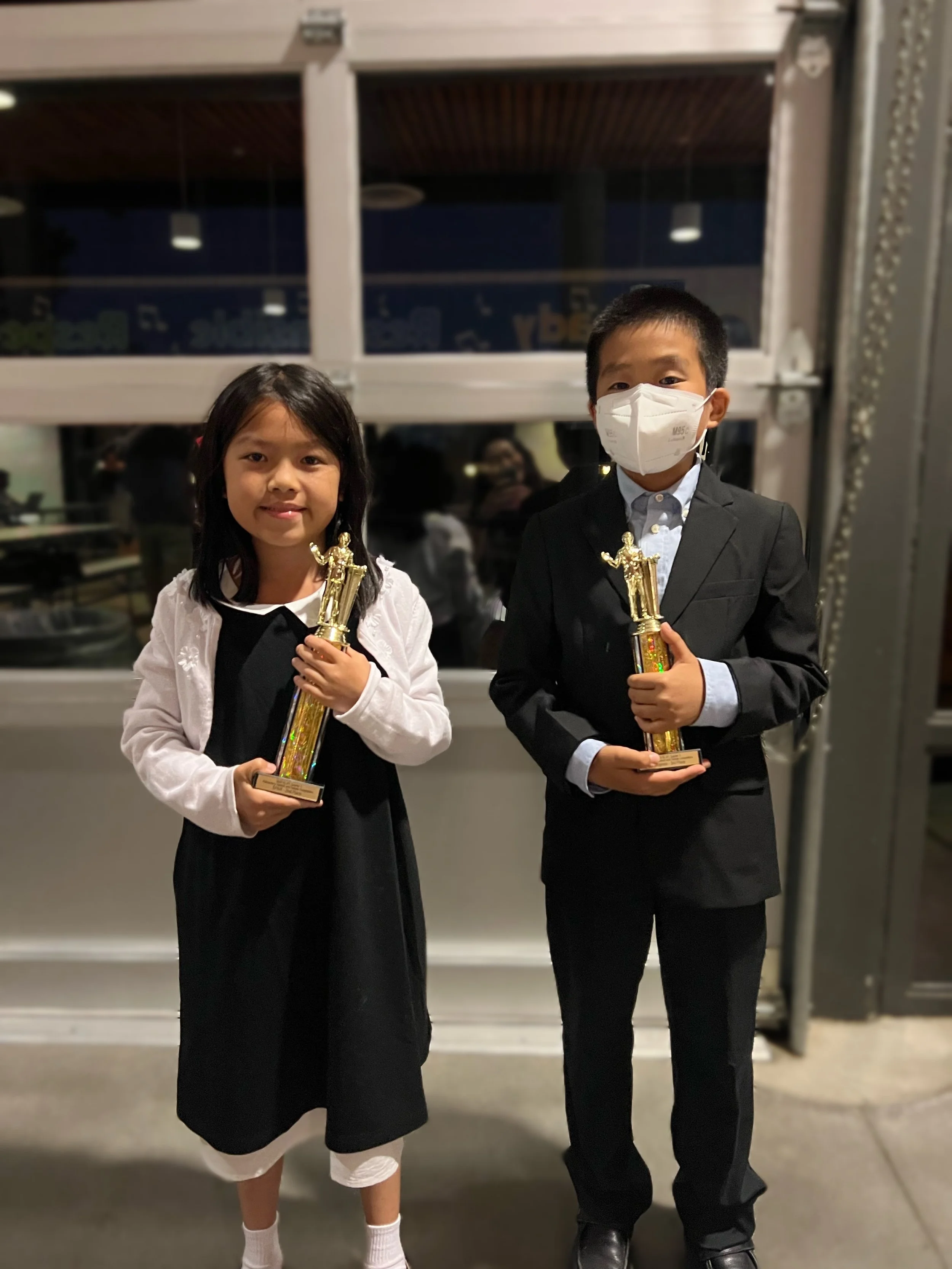 A young girl and boy dressed in formal attire holding trophies, standing in front of a window at night.