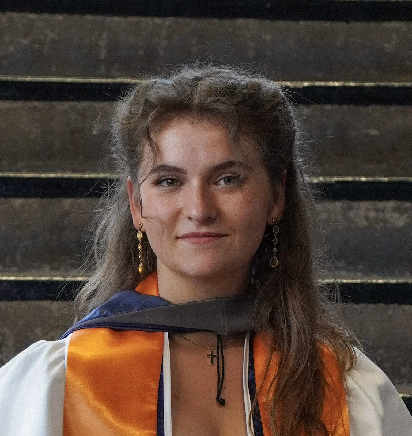 A young woman with long wavy brown hair and earrings, wearing a graduation gown and an orange stole, standing in front of concrete stairs.