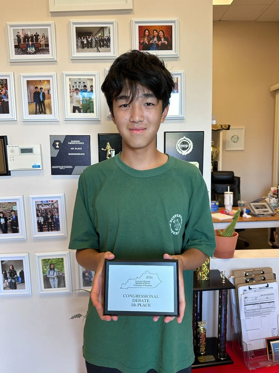 Young man holding an award plaque that reads "Congressional Debate 5th Place" in an office room with framed photos on the wall behind him.
