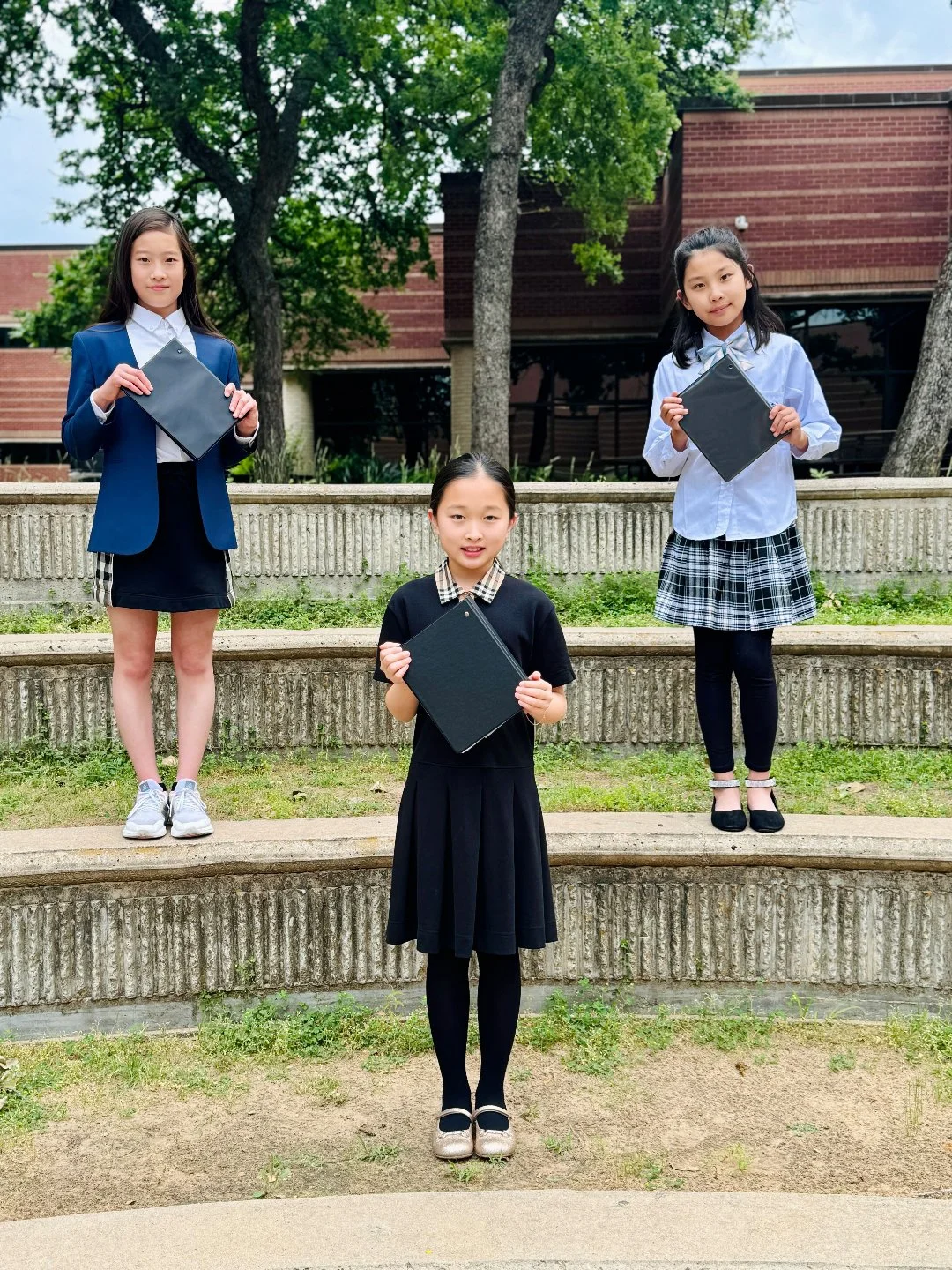 Three young girls in school uniforms standing on steps outdoors, holding black folders, in front of a brick building with trees.