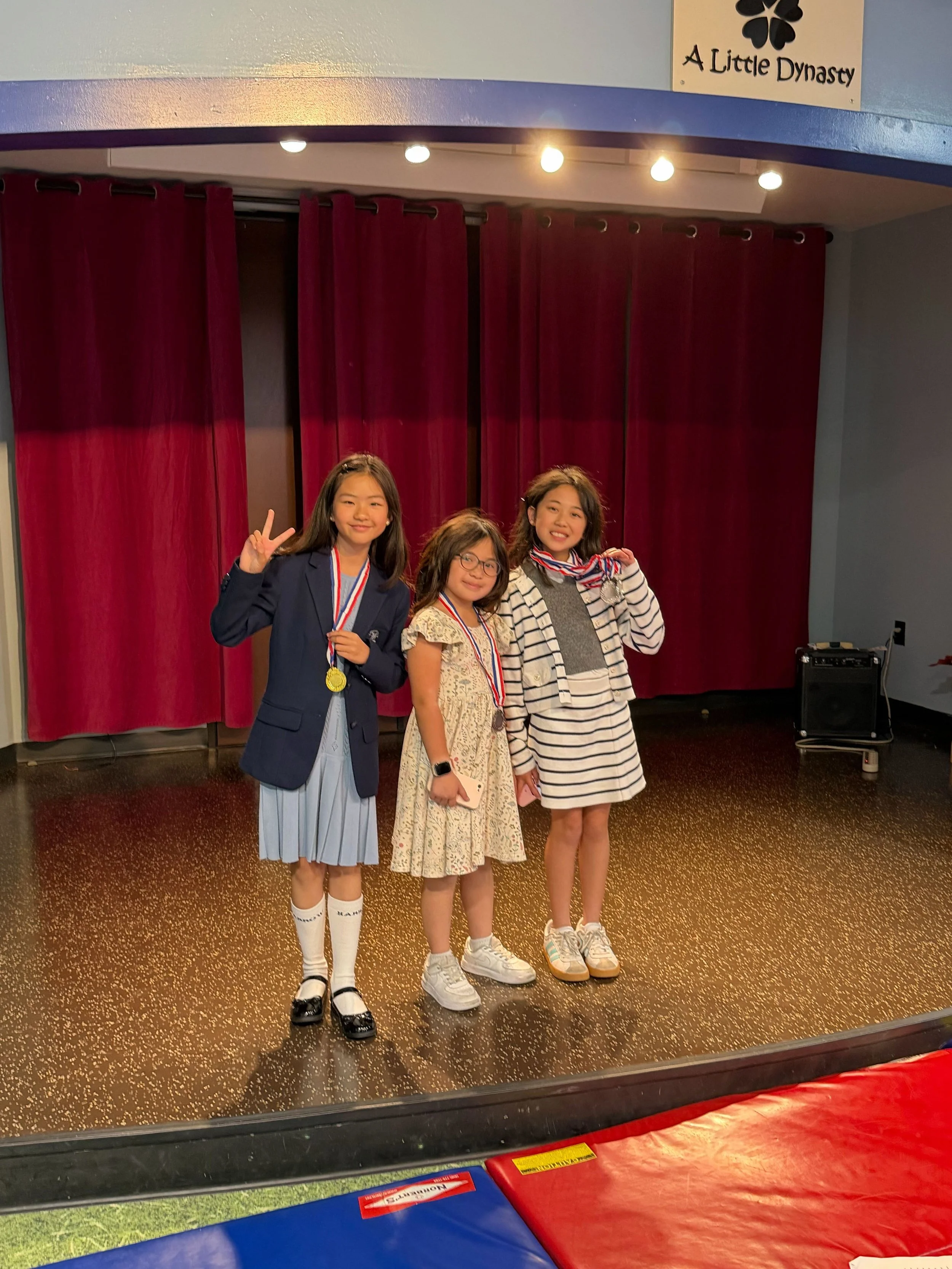 Three young girls standing on a stage, posing and smiling, each wearing medals around their necks, with a red curtain behind them and a sign that says "A Little Dynasty" with a four-leaf clover symbol.