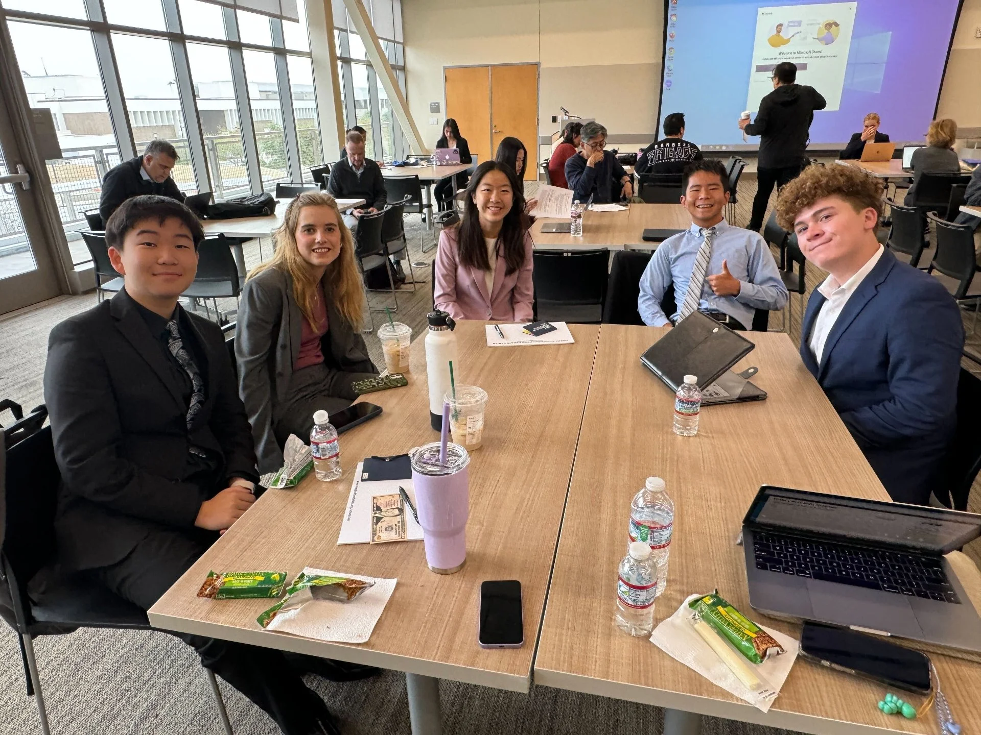 Group of five diverse young professionals in formal attire sitting around a conference table with drinks, snacks, and laptops, in a bright modern conference room with large windows and a presentation screen.