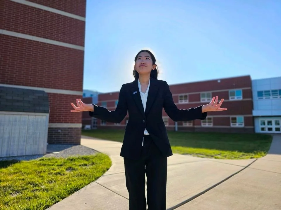A woman in a business suit standing outside on a sidewalk near a brick building, with her arms outstretched and looking up at the sky.