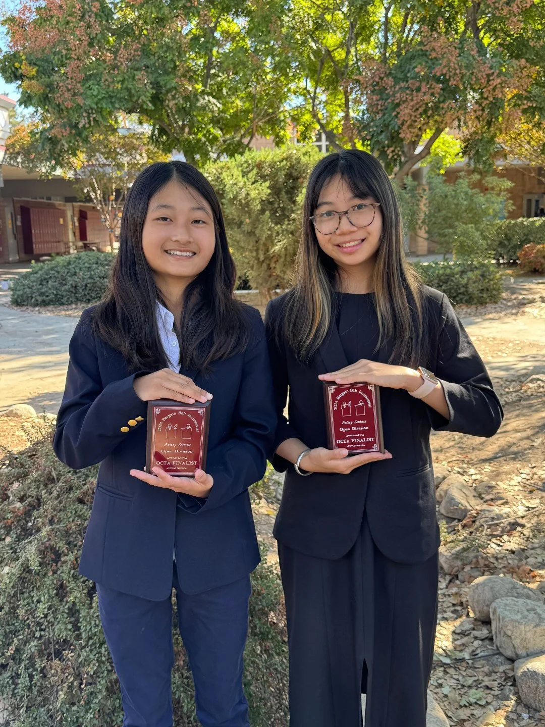 Two young women in business suits holding awards outside in front of trees and bushes.
