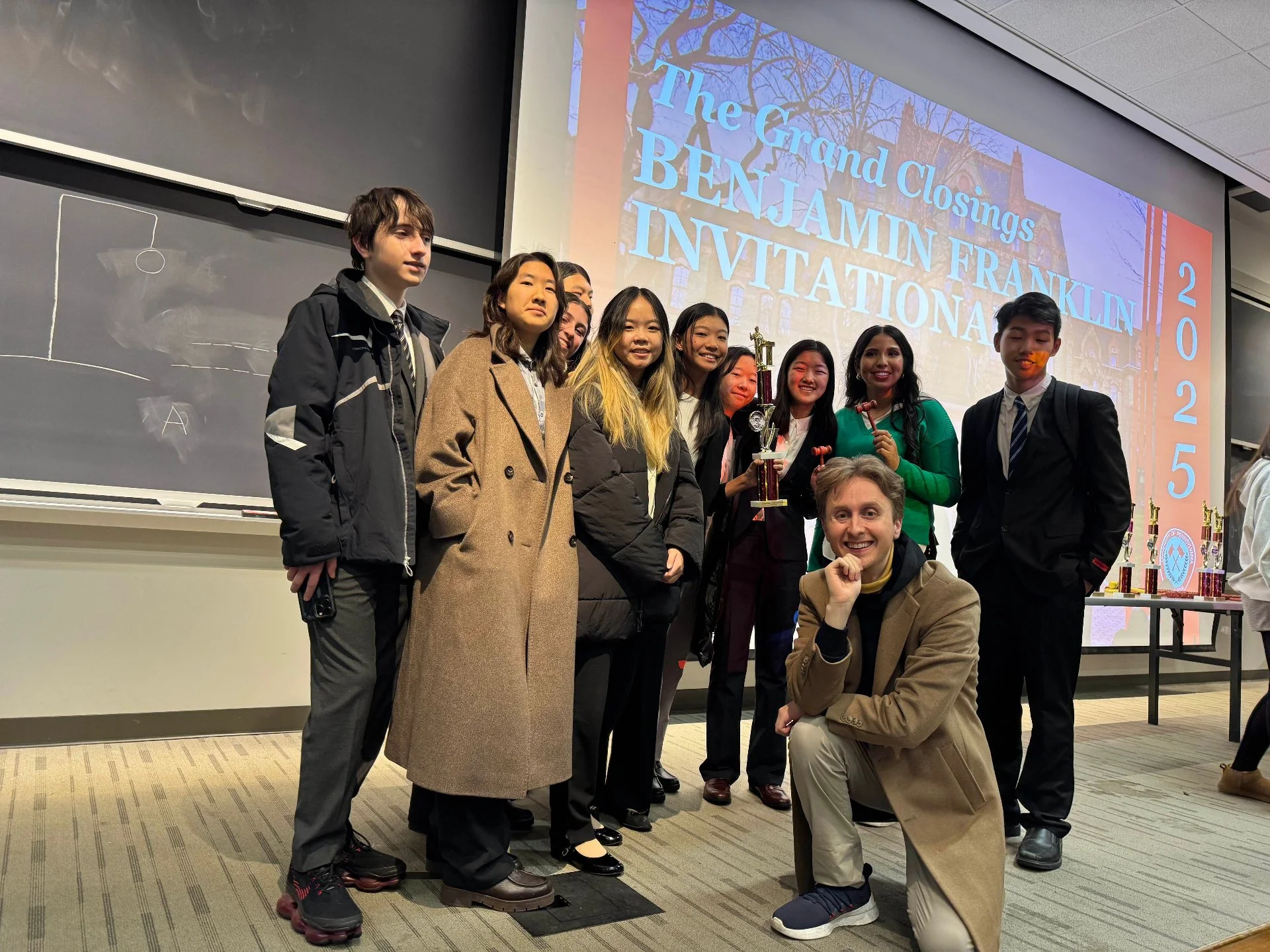 Group of students and a teacher holding trophies in a classroom during an awards ceremony, with a presentation slide in the background reading 'The Grand Closings Benjamin Franklin Invitational 2025'.