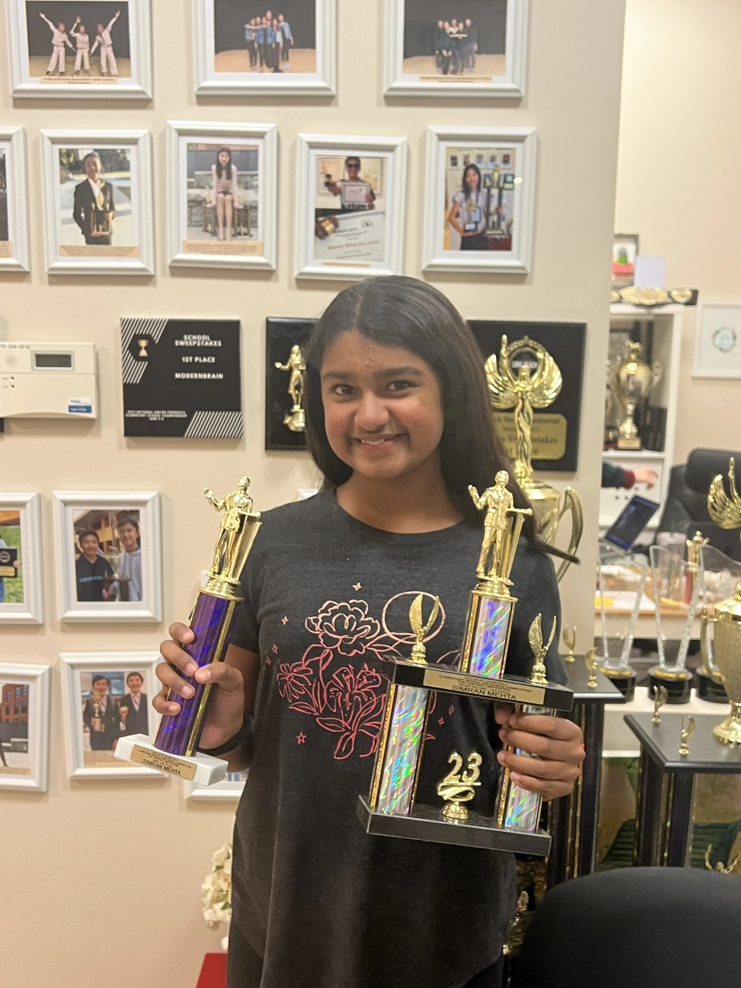 A young girl smiling and holding two trophies in a room decorated with framed photographs and awards on the wall behind her.