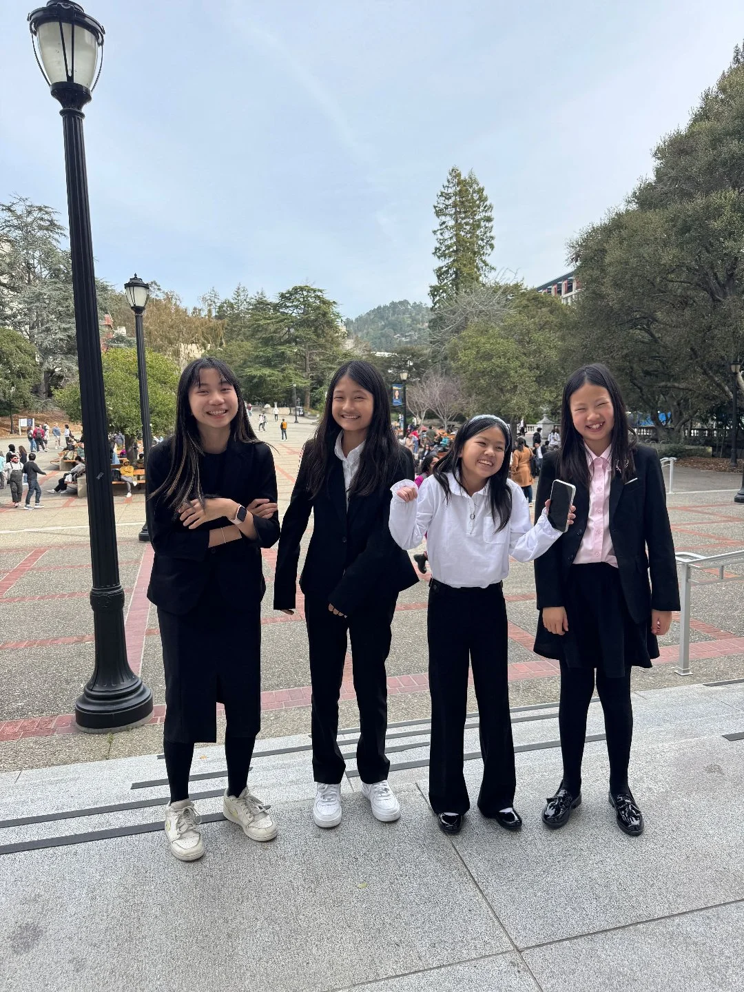 Four young women dressed in business casual attire standing outdoors, smiling, with trees and people in the background.