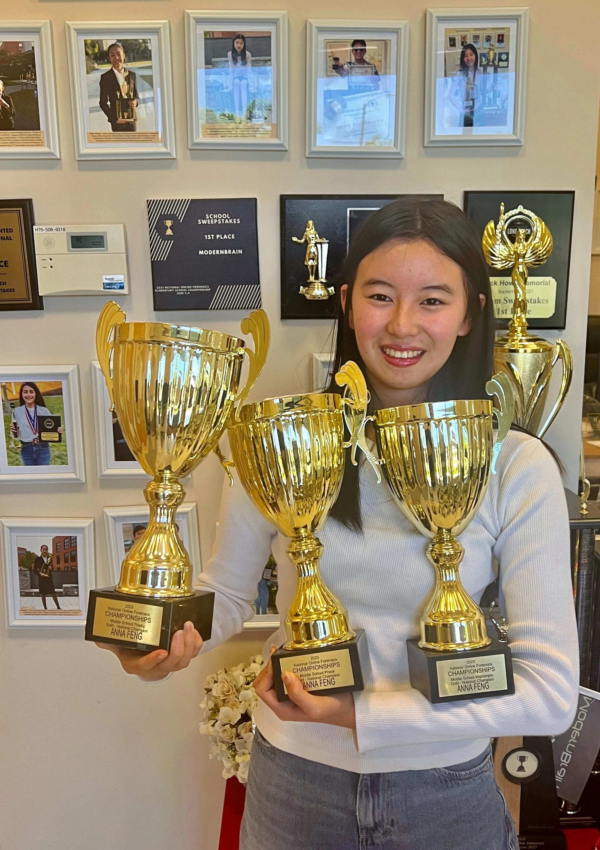 A young woman holding three large gold trophies, smiling, standing in front of a wall decorated with framed photos and awards.