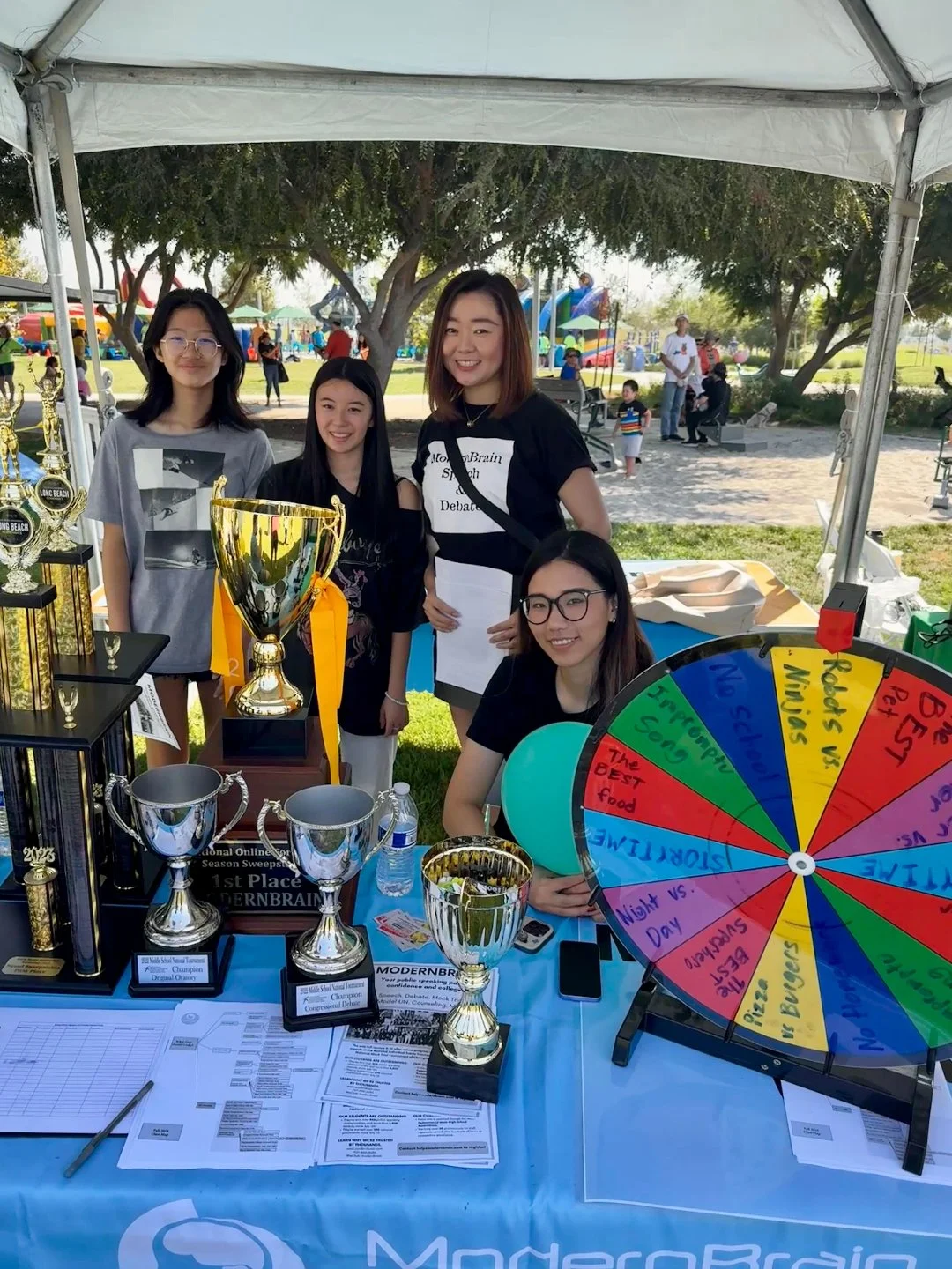A group of five young women at an outdoor event with trophies, a spin wheel, and informational materials on a table, under a canopy with trees and playground in the background.