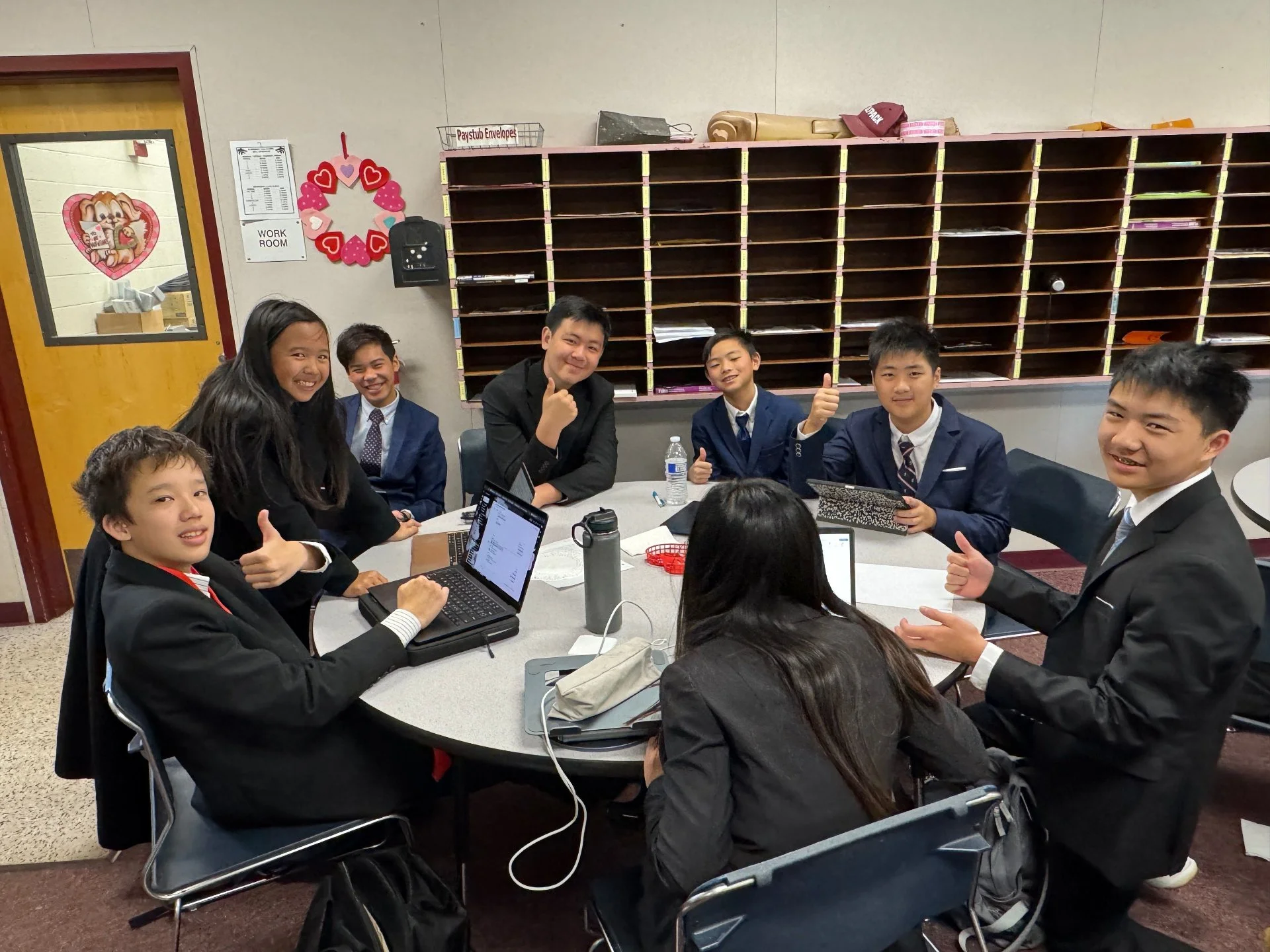 Group of seven students and one adult in formal attire sitting around a table in a school classroom, smiling, with some giving thumbs up. The table has laptops, notebooks, and a water bottle. The classroom has a wooden shelf with cubby holes on the w