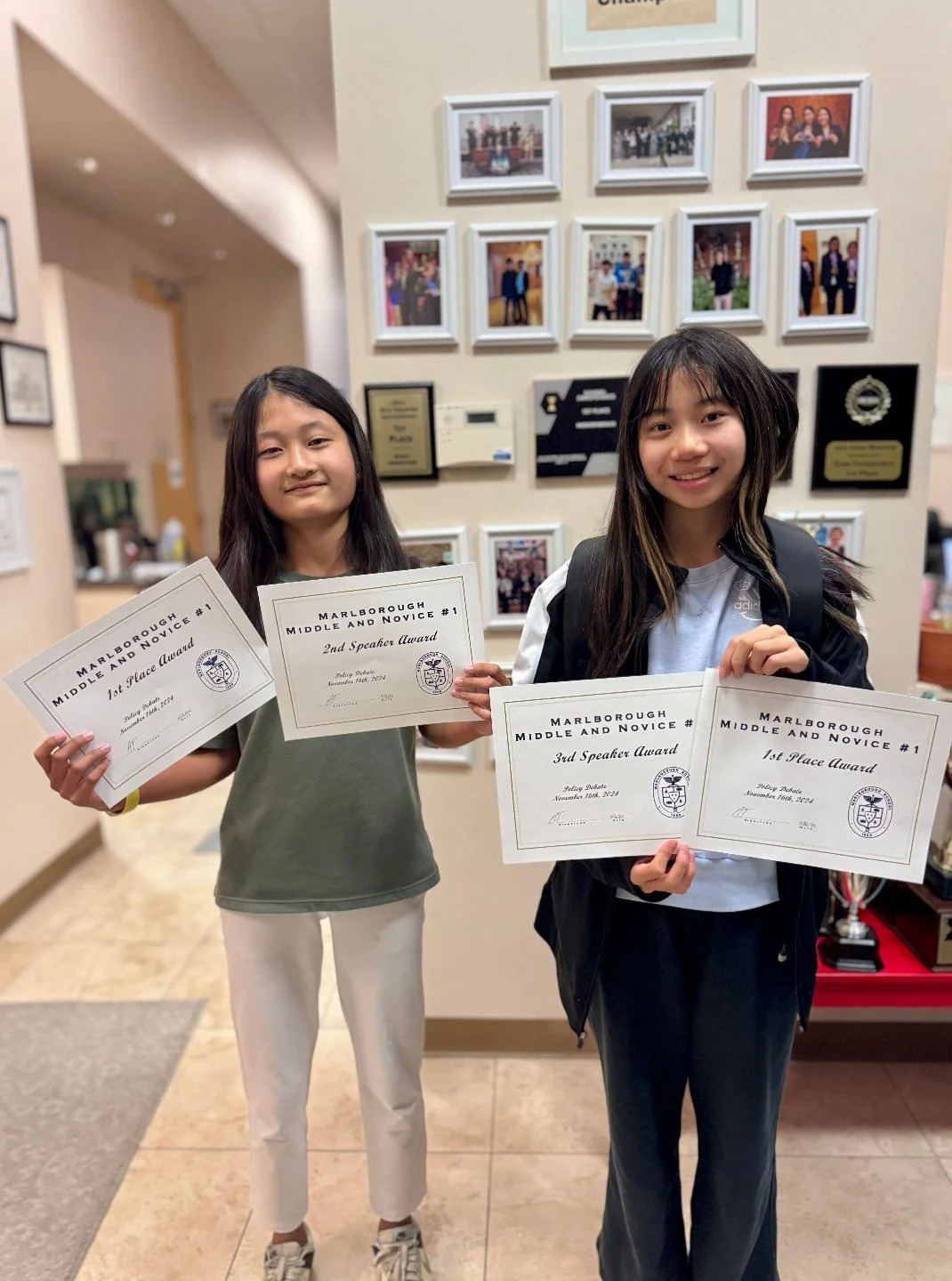 Two girls standing inside a school building holding certificates for academic achievement. The girl on the left has long dark hair, wears a green shirt and white pants, and holds certificates for first and second place. The girl on the right has long