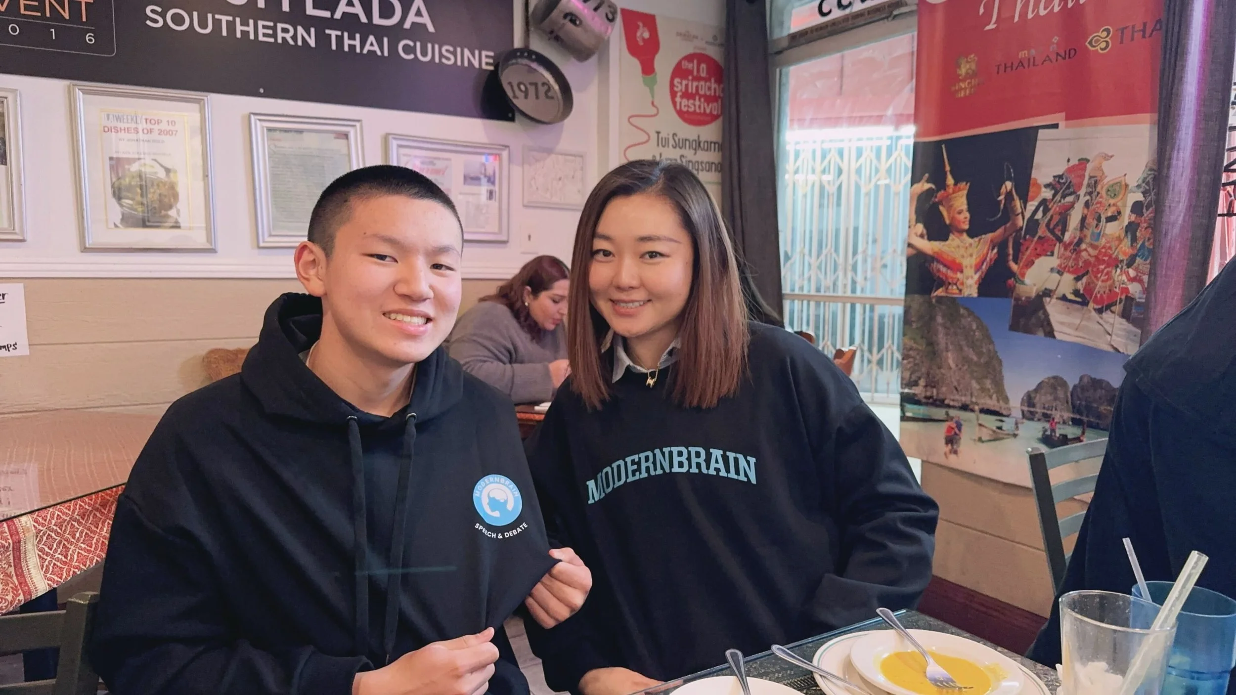 Two young people sitting at a table inside a Thai restaurant, smiling at the camera. The person on the left is holding a black hoodie with a logo, and the person on the right is wearing a black shirt with white text. Behind them, there are framed pic