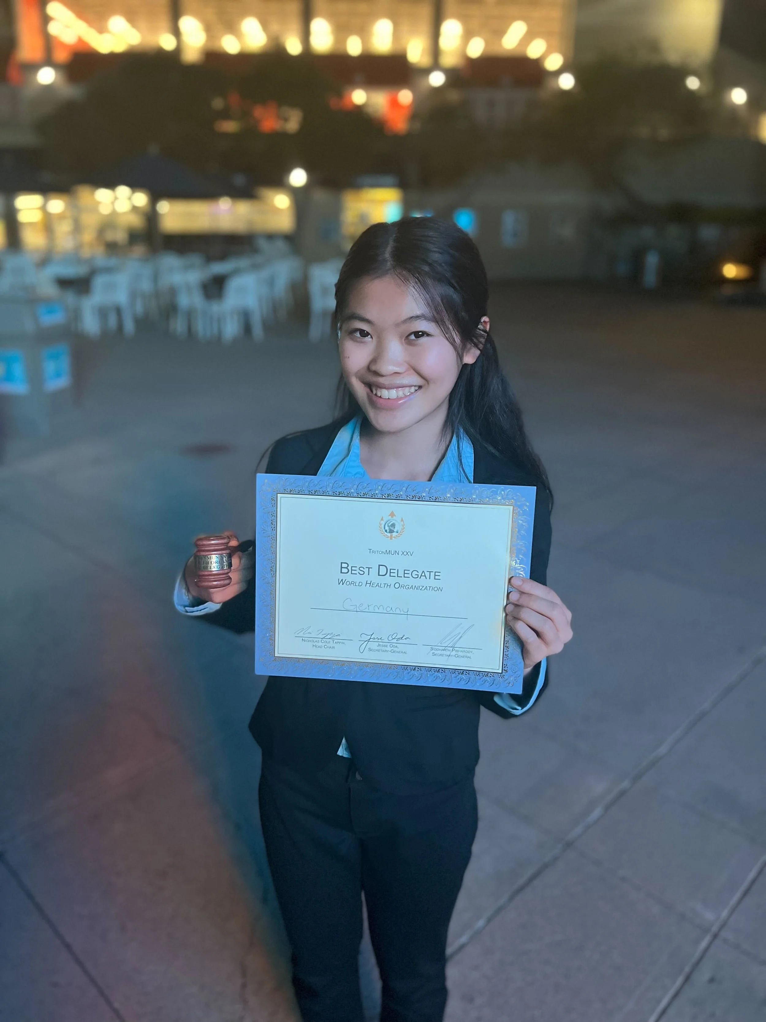 A young woman in formal attire smiling and holding a certificate that reads 'Best Delegate, World Health Organization' and 'Germany.'