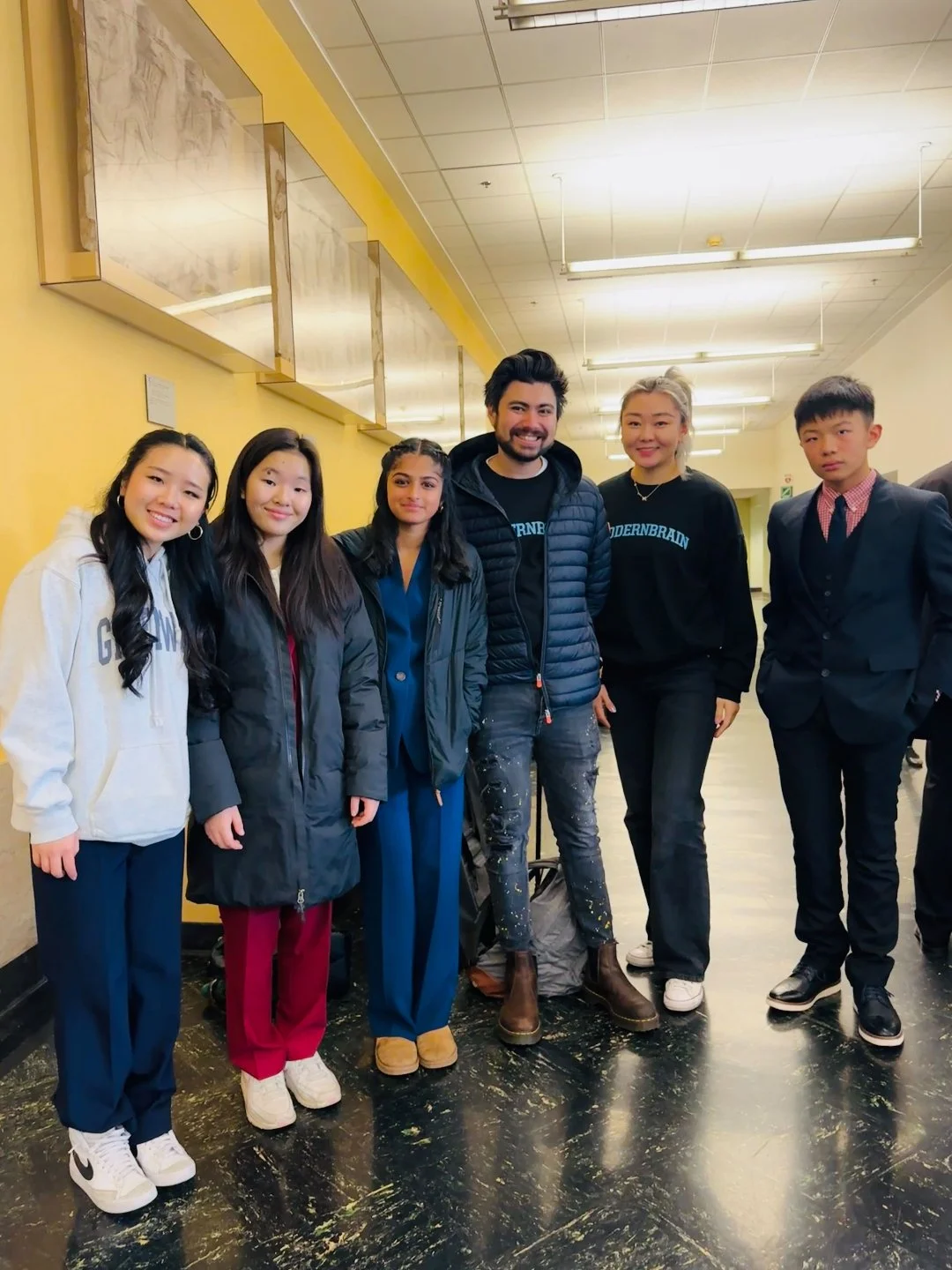 Group photo of six young people standing together in an indoor hallway, smiling and posing for the camera.