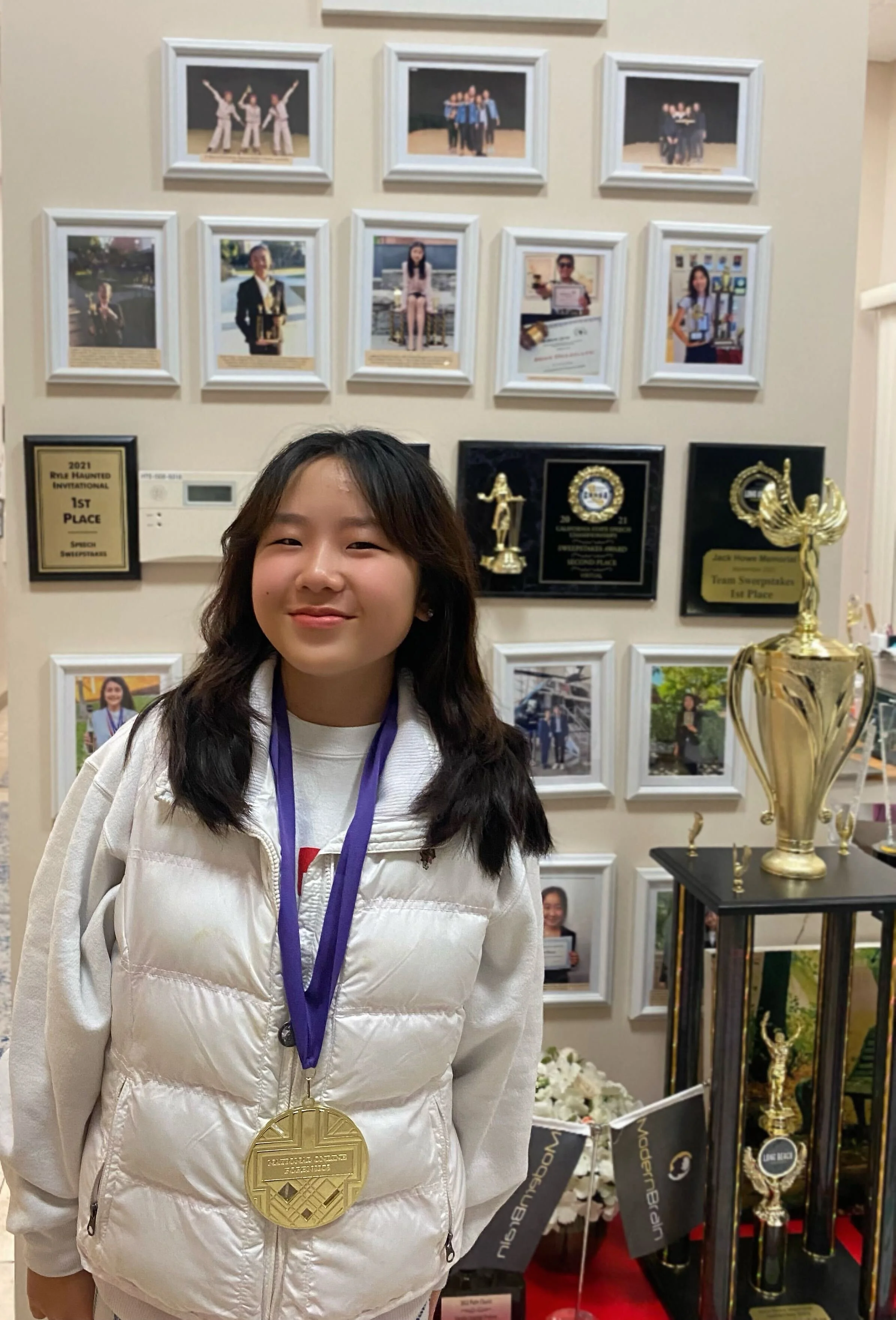 A young girl with long dark hair wearing a white puffy vest and a gold medal around her neck, standing in front of a wall with framed photographs and awards.