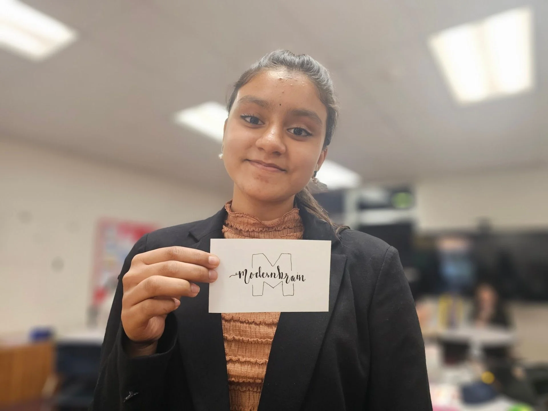 A young woman in a black blazer holding a white card with handwritten 'Modernrain' and a sketch of the letter ‘M’ in the background, standing in an indoor setting.