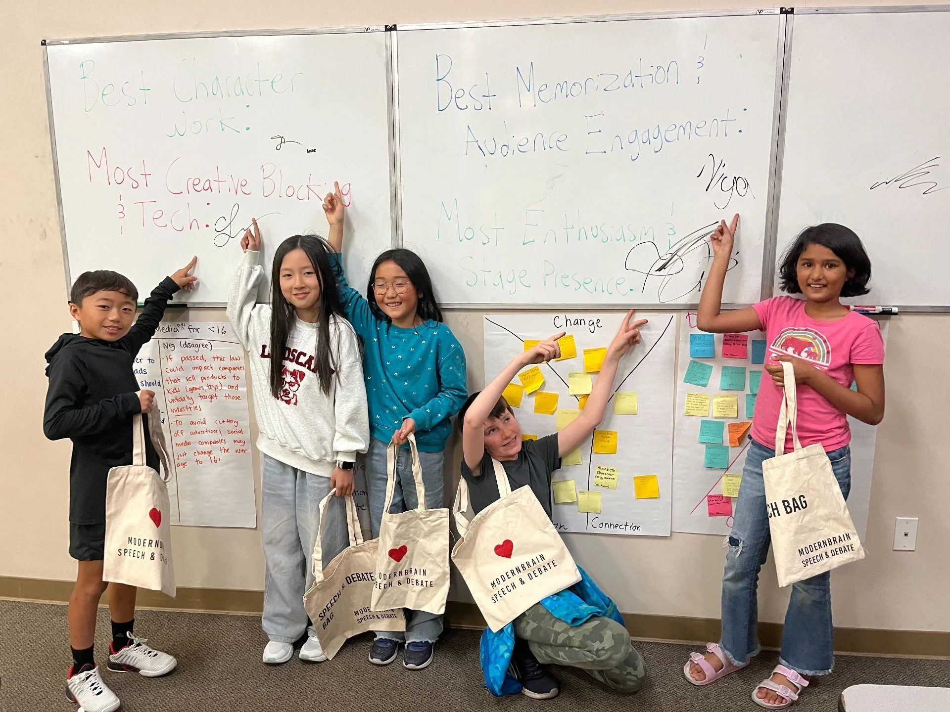 Group of five children standing in front of whiteboards with notes and sticky notes, holding tote bags from a speech and debate event, smiling and pointing at the boards.