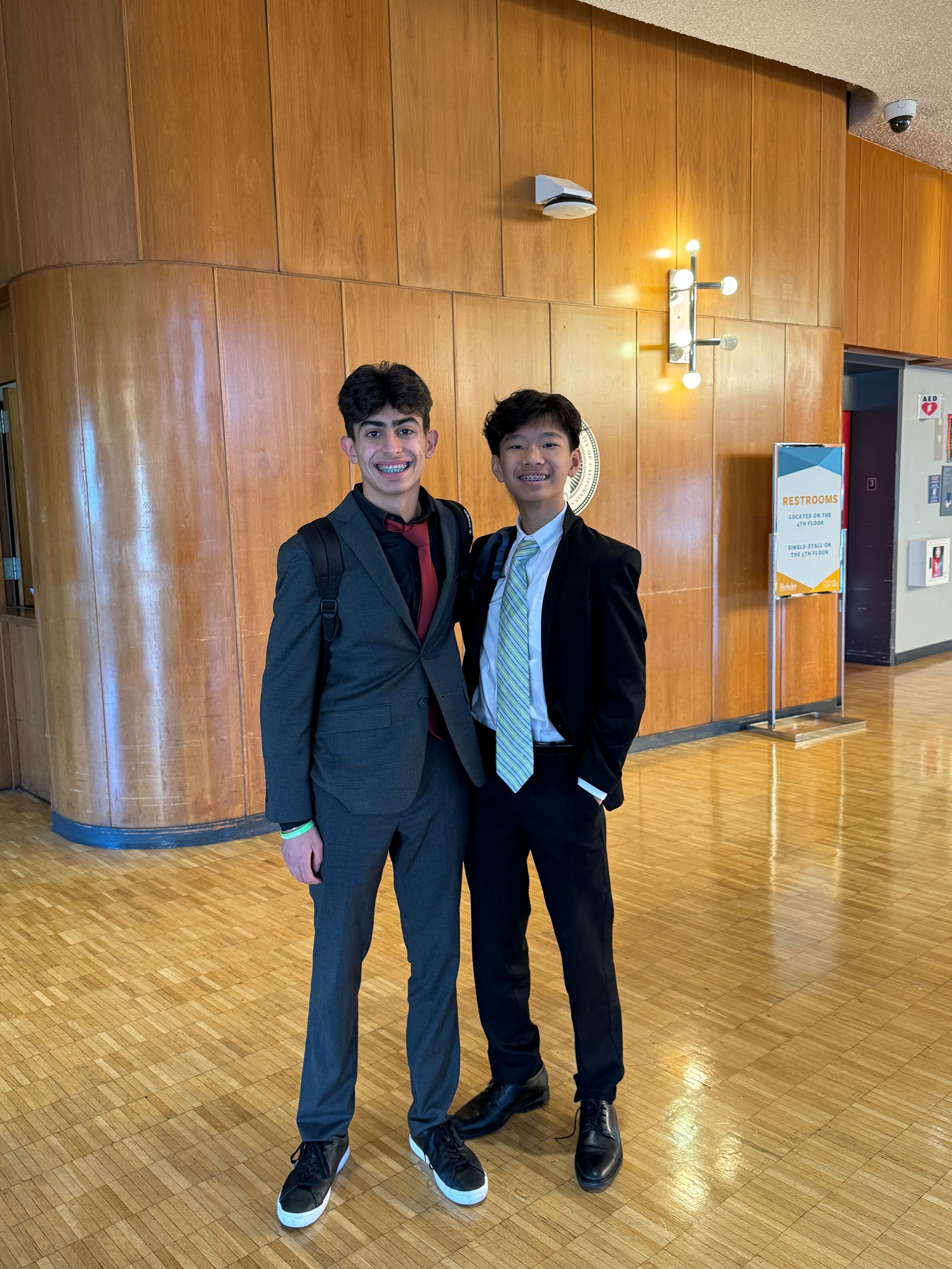 Two young men in suits and backpacks standing together in a wooden-floored hall, smiling at the camera.