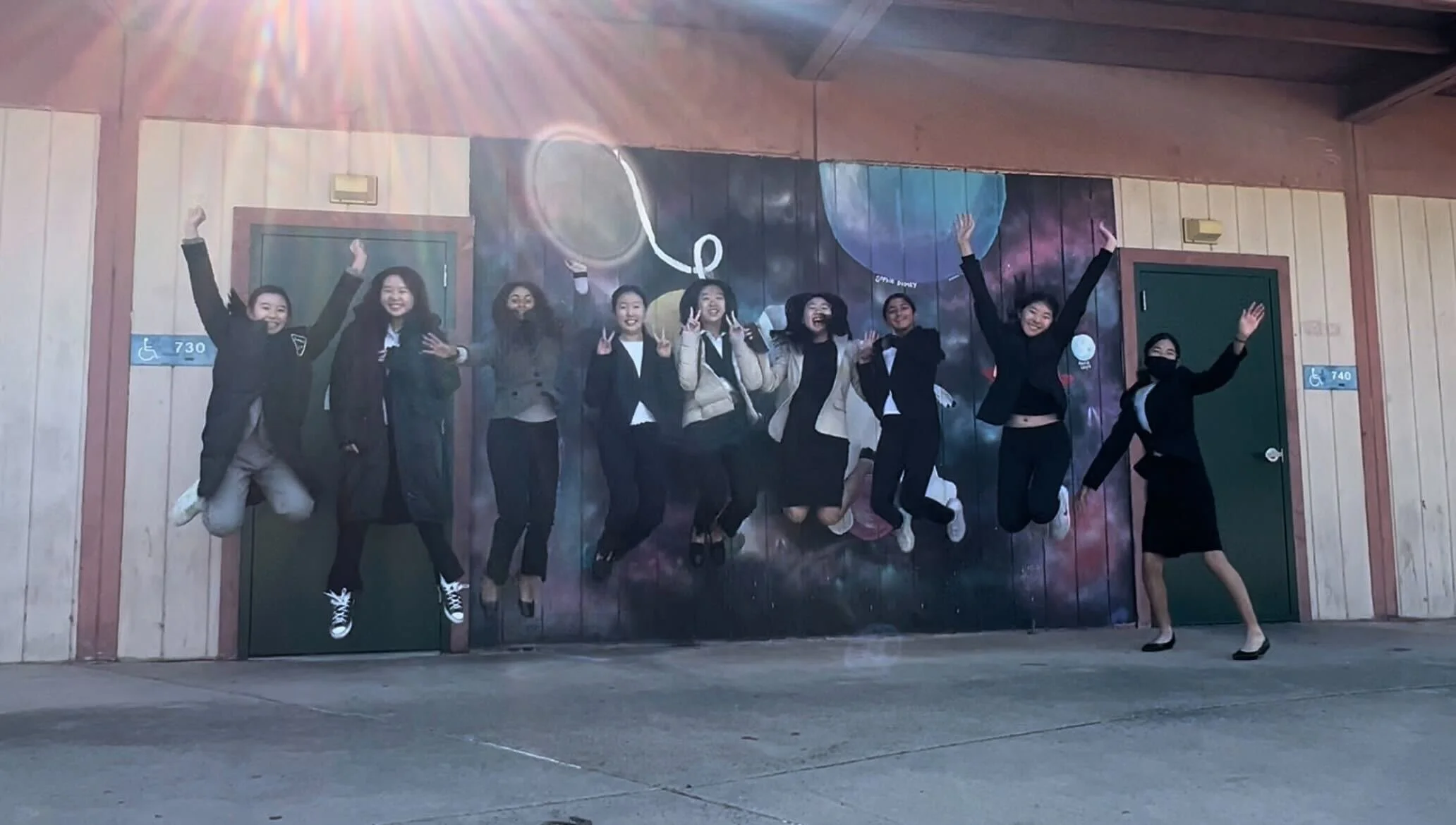 Group of eight young women posing and jumping in front of a colorful space-themed mural at a building with wooden panels, two of the women are wearing black masks, and they are in a room with concrete floor.