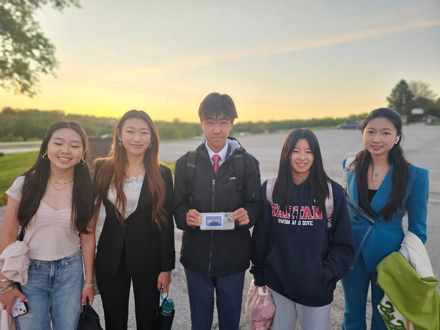 Five young people standing outdoors during sunset, smiling and dressed casually, with some holding bags and a phone.