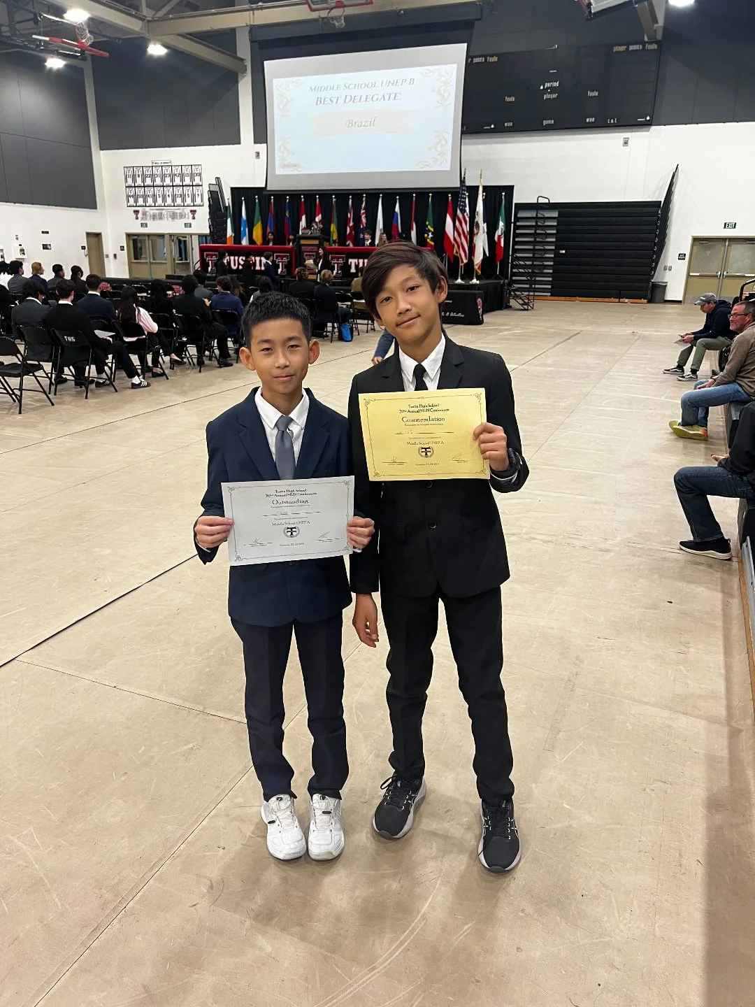 Two boys in suits holding certificates at an indoor award ceremony with flags and a stage in the background.