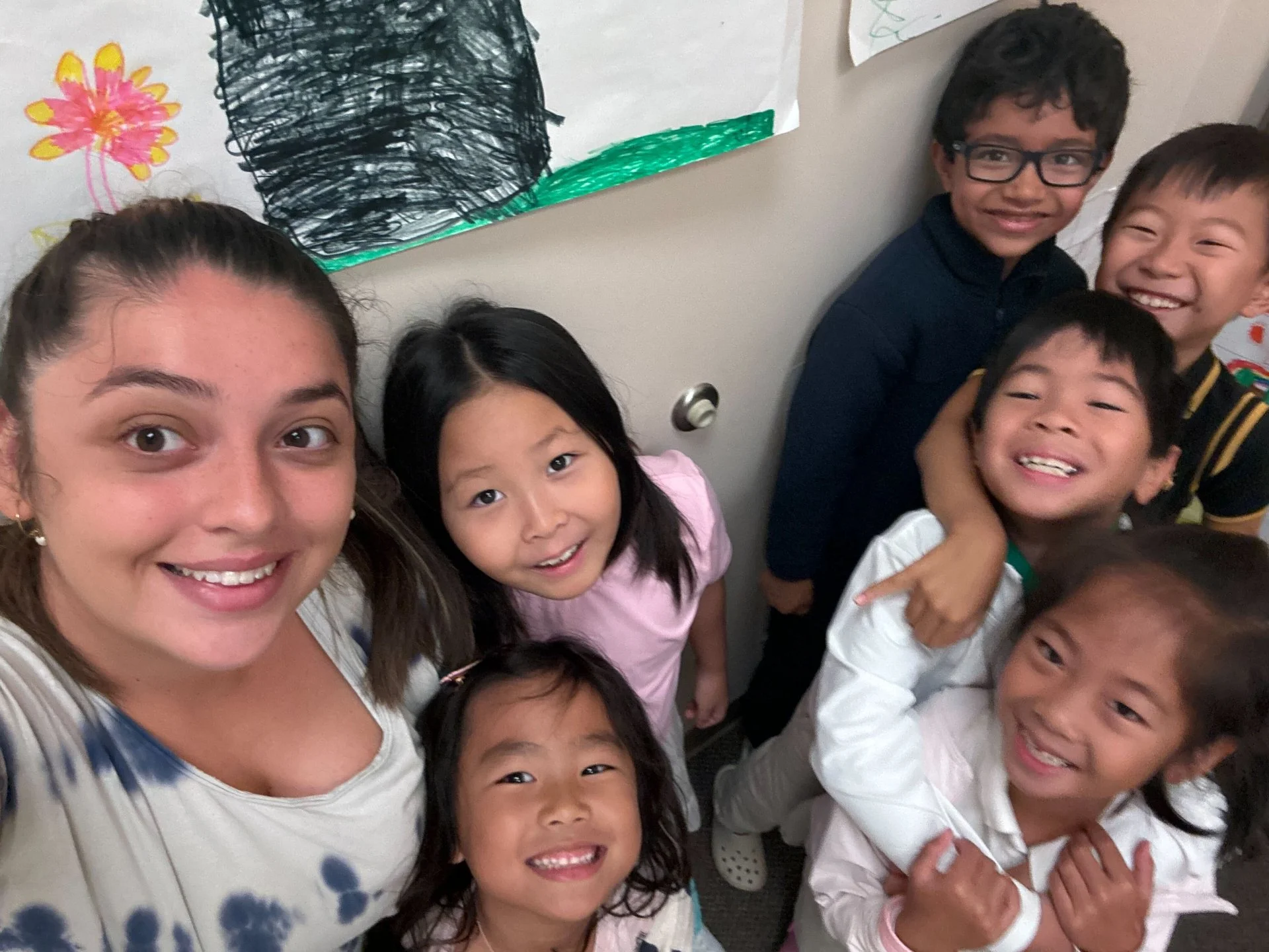 A smiling woman with six children, posing in front of colorful drawings on the wall.