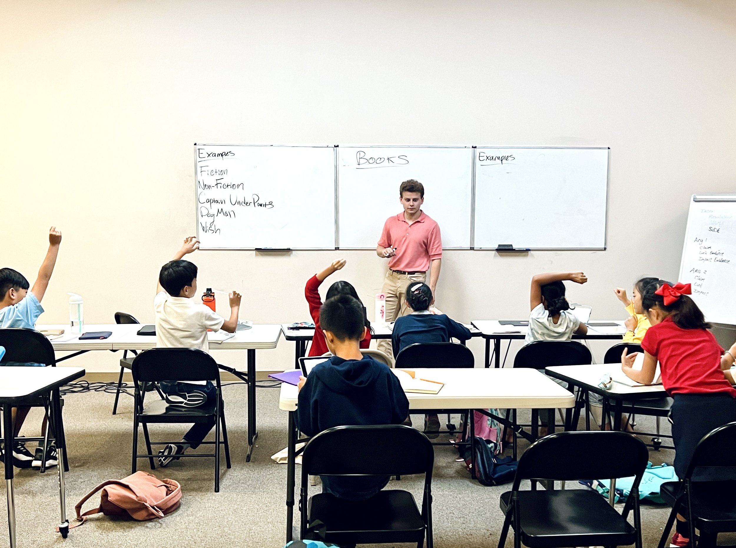 A classroom with young students sitting at desks, raising their hands, and a teacher standing in front of a whiteboard.