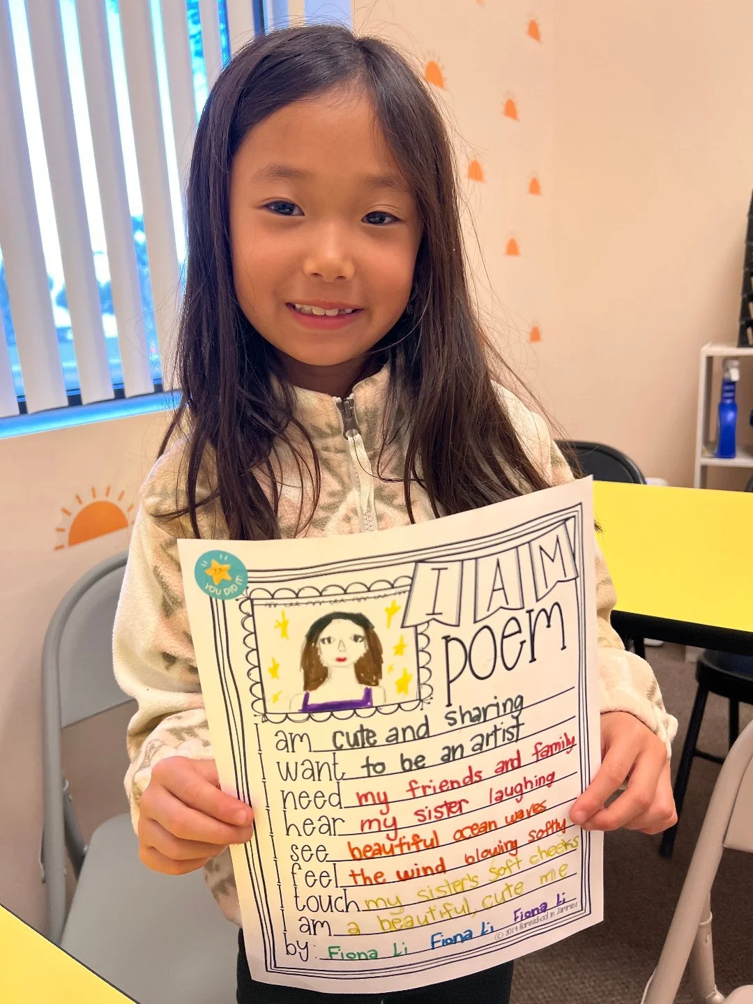 A young girl with long dark hair holding a colorful handwritten poem with a drawing of herself on white paper. She is smiling and standing in a classroom with orange and blue decorations in the background.