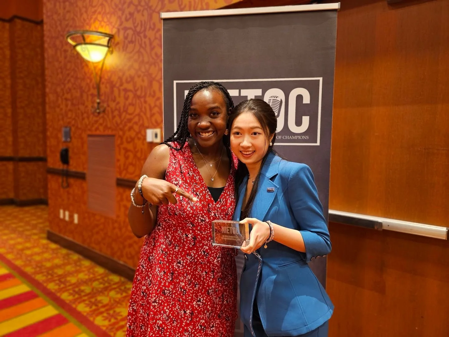 Two women standing together at an event, one holding an award, in front of a black banner with the word "METOO." The woman on the left has dark skin, is wearing a red patterned sleeveless dress, and is pointing at the award. The woman on the right ha