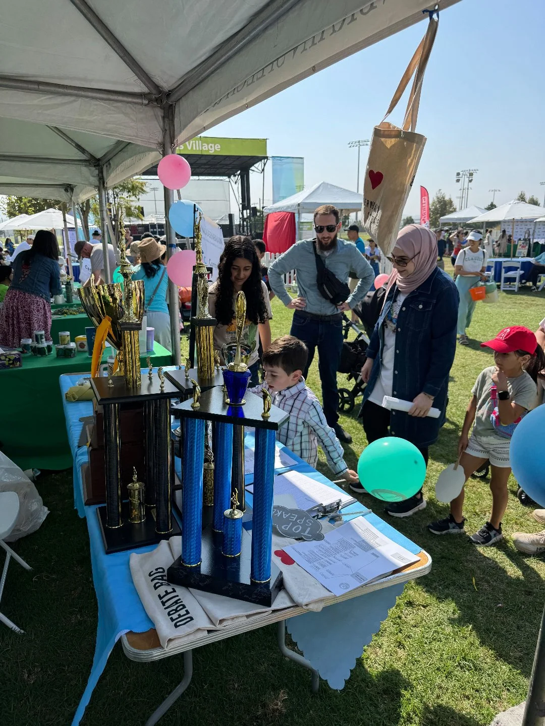 At an outdoor fair, a table displays trophies, balloons, and event materials under a canopy. People, including children and adults, are gathered around, engaging with the display and each other.