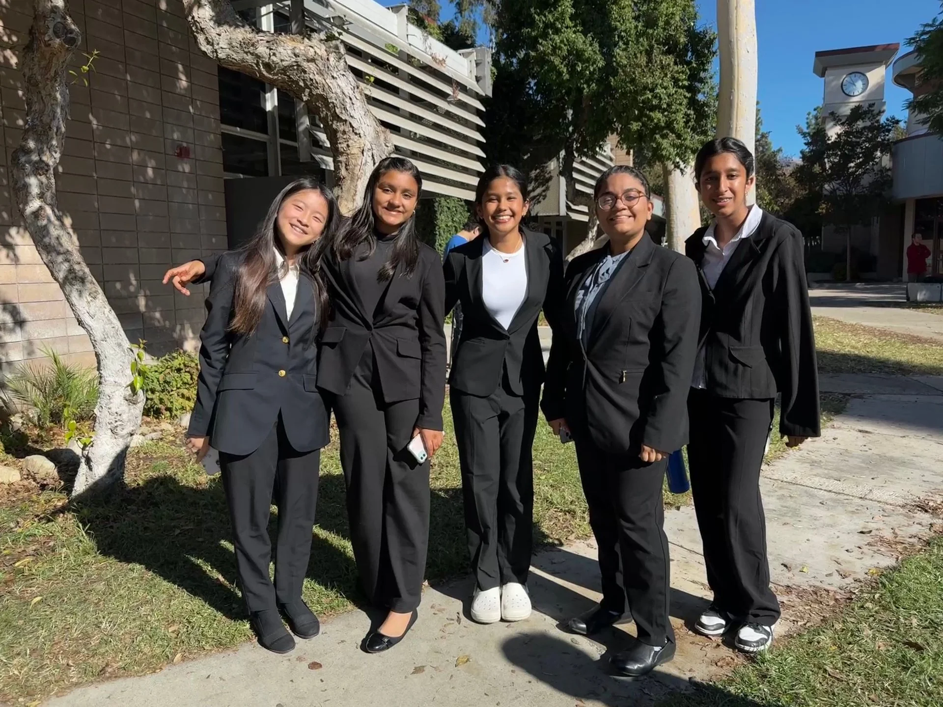 Five young women in formal black suits standing outdoors on a sunny day, smiling for the camera.