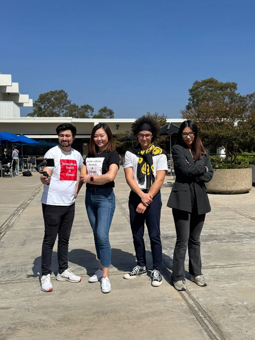 Four people standing outdoors on a concrete surface, smiling and posing for the photo, with a building, trees, and a blue sky in the background.