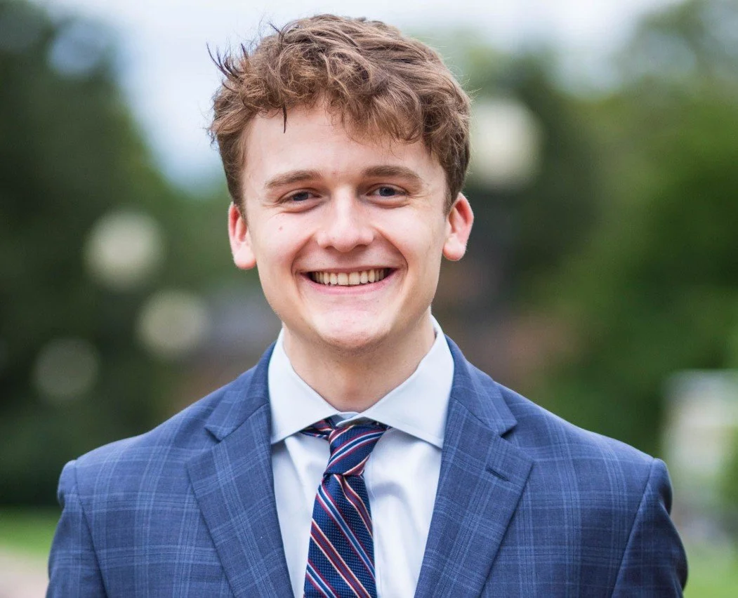 Young man in a blue checkered suit and striped tie smiling outdoors with blurred greenery in the background.