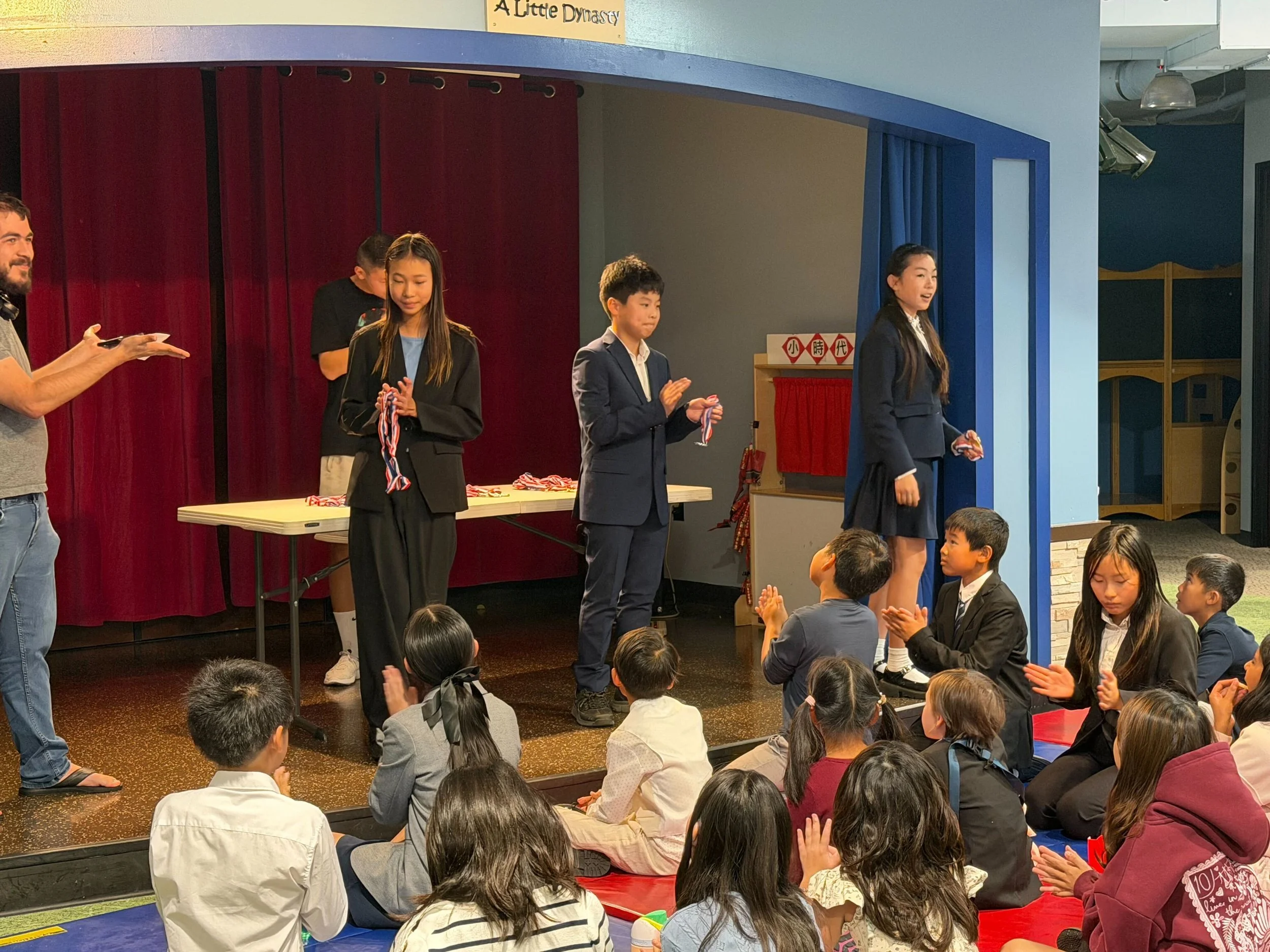 Kids receiving medals during an award ceremony on a stage with an audience of children sitting on the floor.