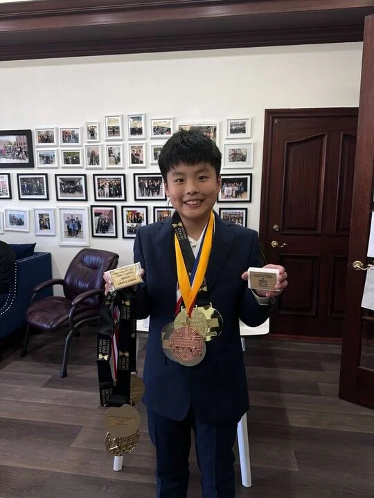 A young boy in a navy suit stands indoors, smiling, wearing multiple medals around his neck, holding two trophies, with a wall of framed photos behind him.