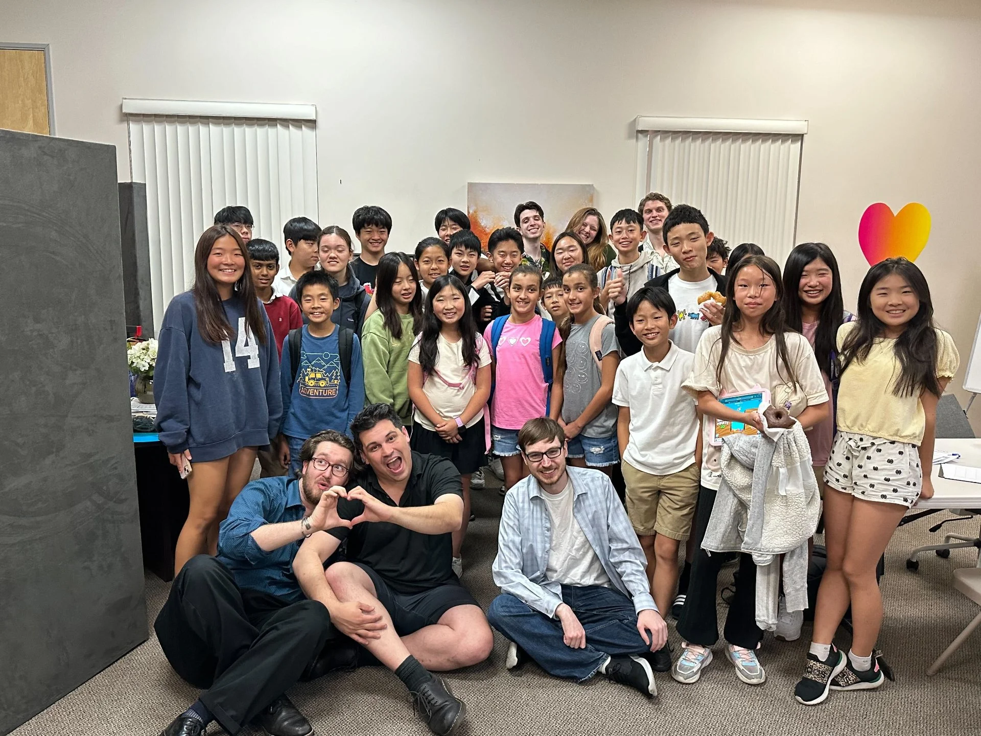 Group photo of smiling children and three adult men in a classroom with two windows, some with backpacks, and a colorful heart-shaped balloon on the wall.