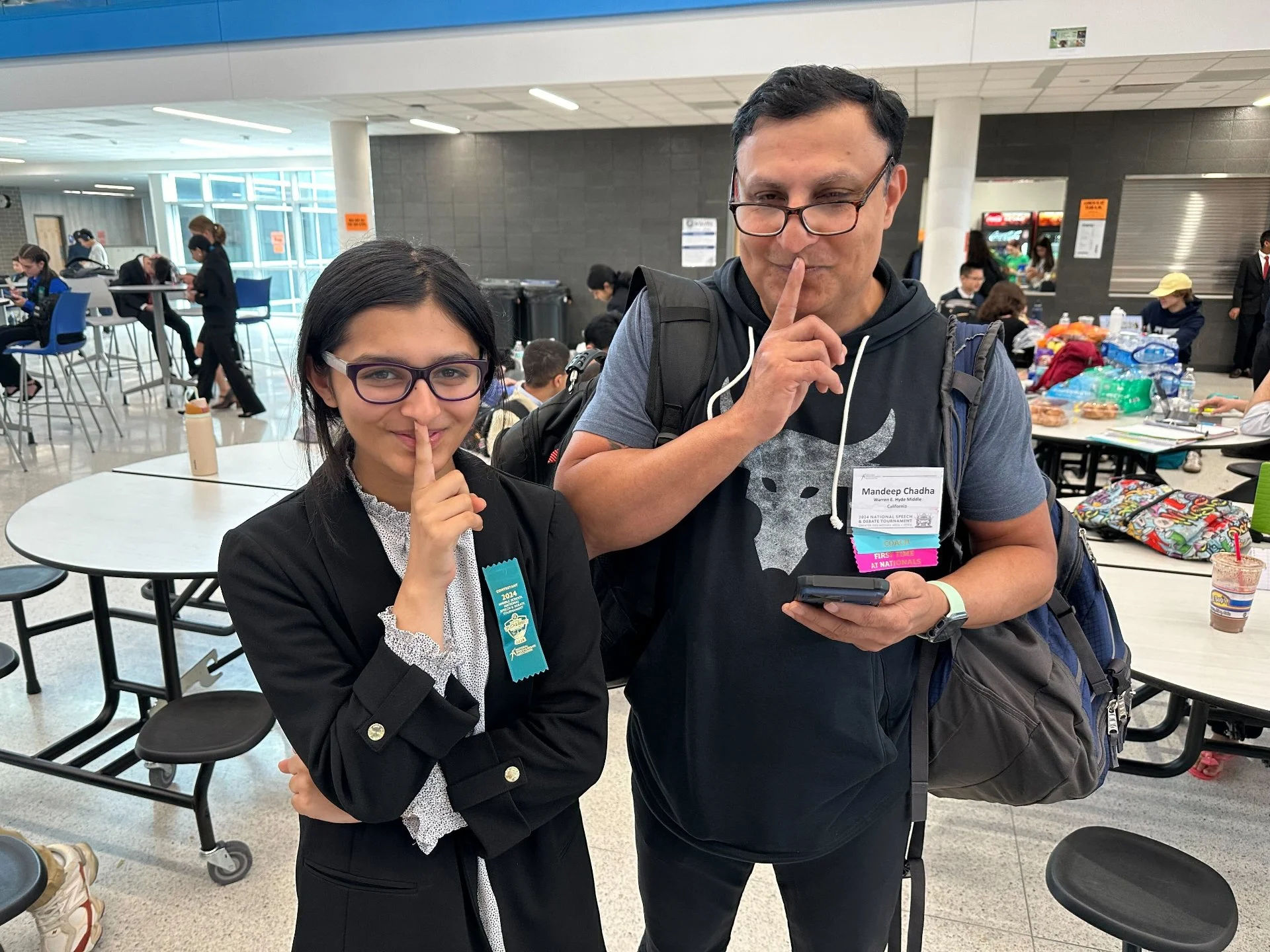 A young girl and an adult man holding a finger to their lips in a 'shh' gesture inside a bustling indoor venue, with tables, chairs, and people in the background.