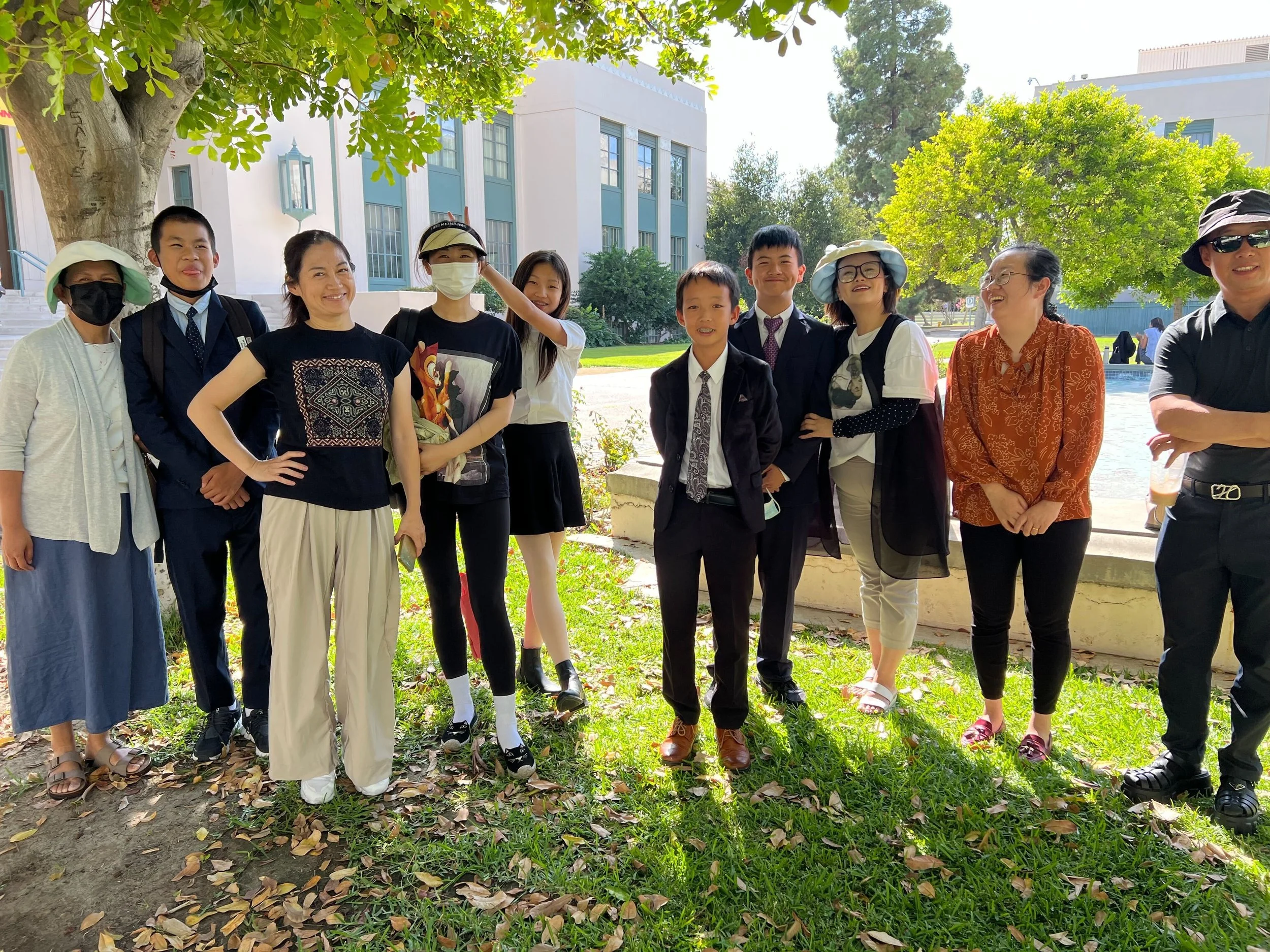 Group of people outdoors on a sunny day, standing under a large tree with green leaves. There are nine women and children dressed in formal and casual clothes, some wearing hats and masks, standing on grass with fallen leaves. Behind them are modern 