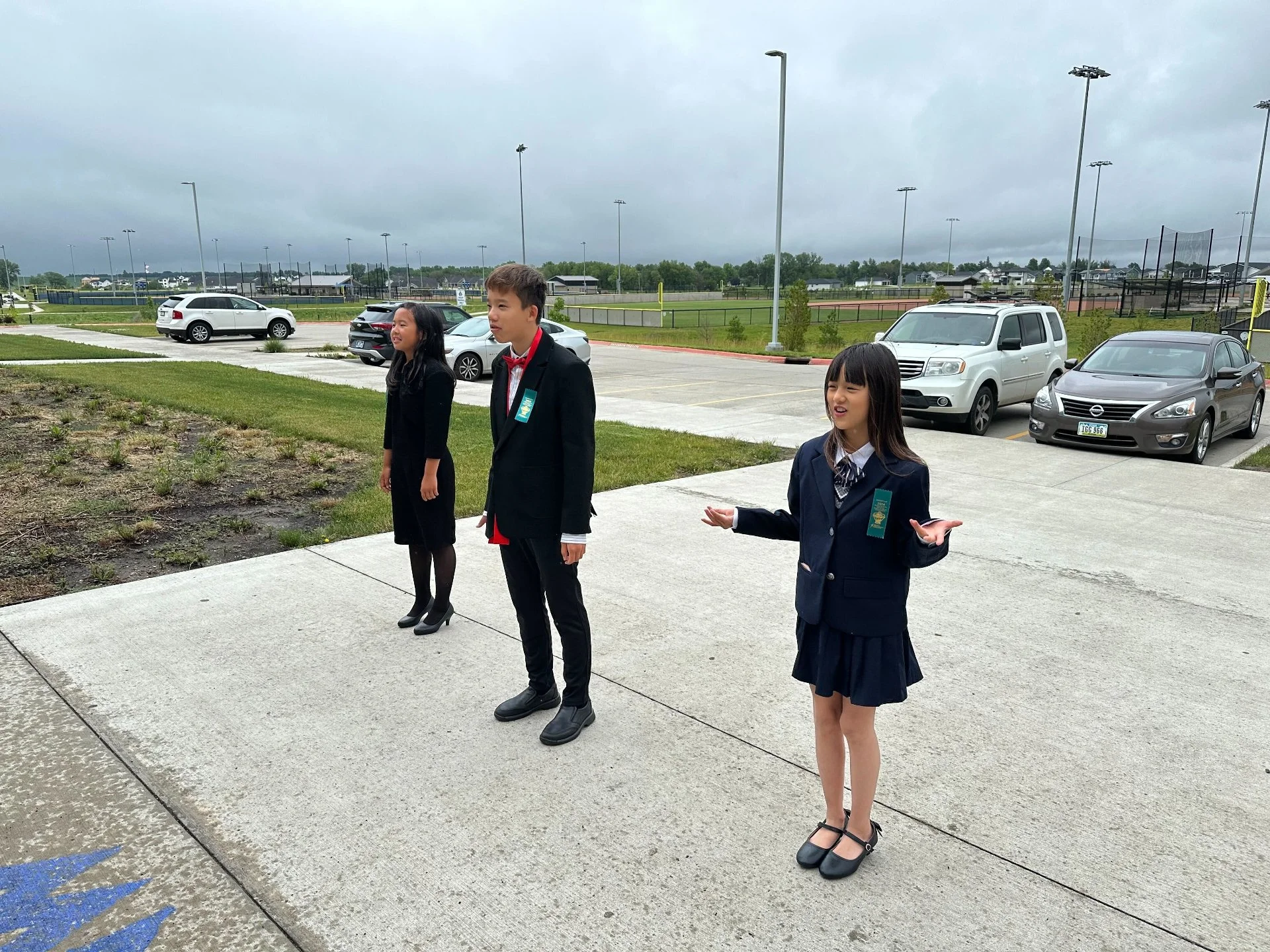 Three children wearing school uniforms standing on a sidewalk outdoors next to parked cars with a sports field in the background, under cloudy skies.